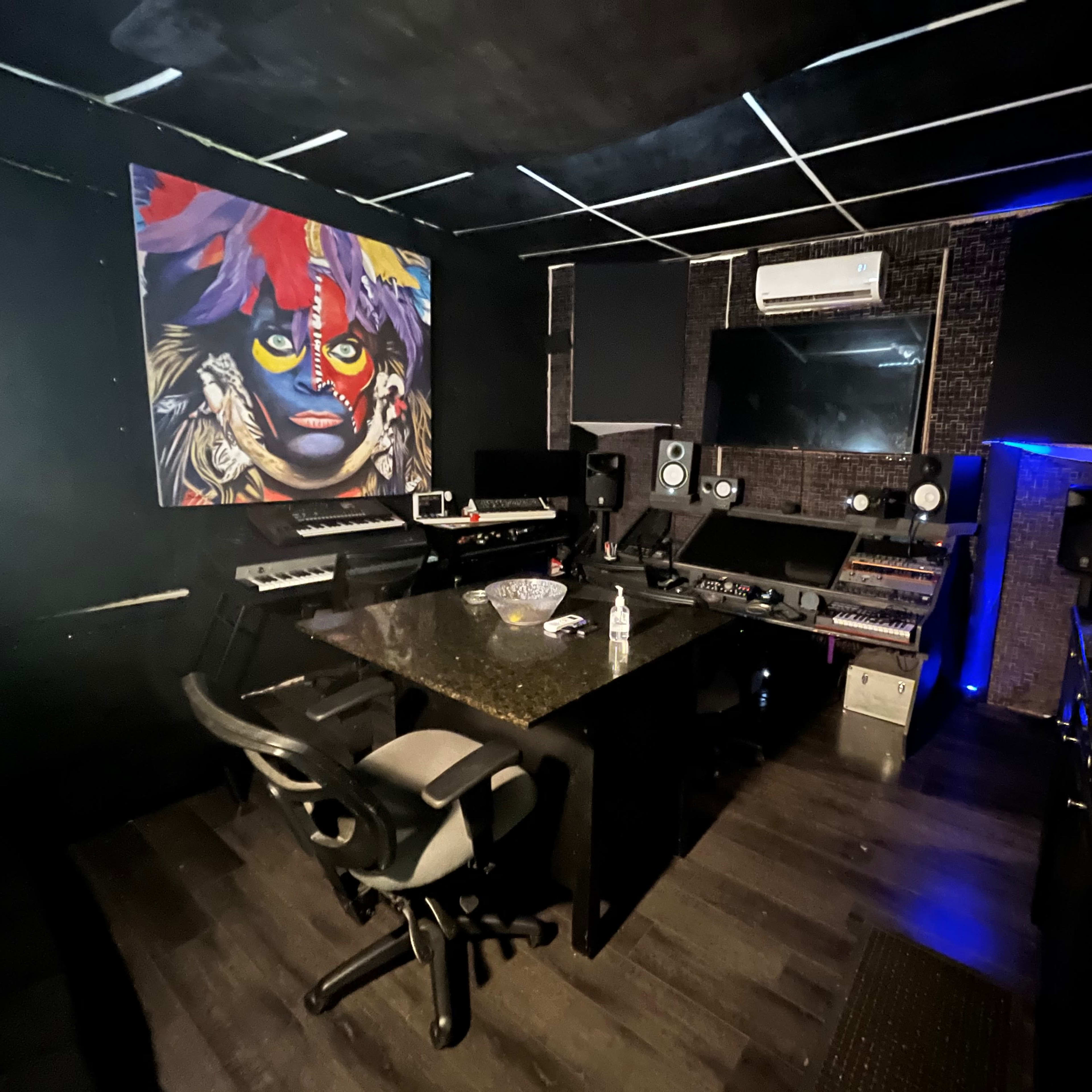 A music studio featuring a central control desk, various electronic instruments, and a large colorful mural on the wall.