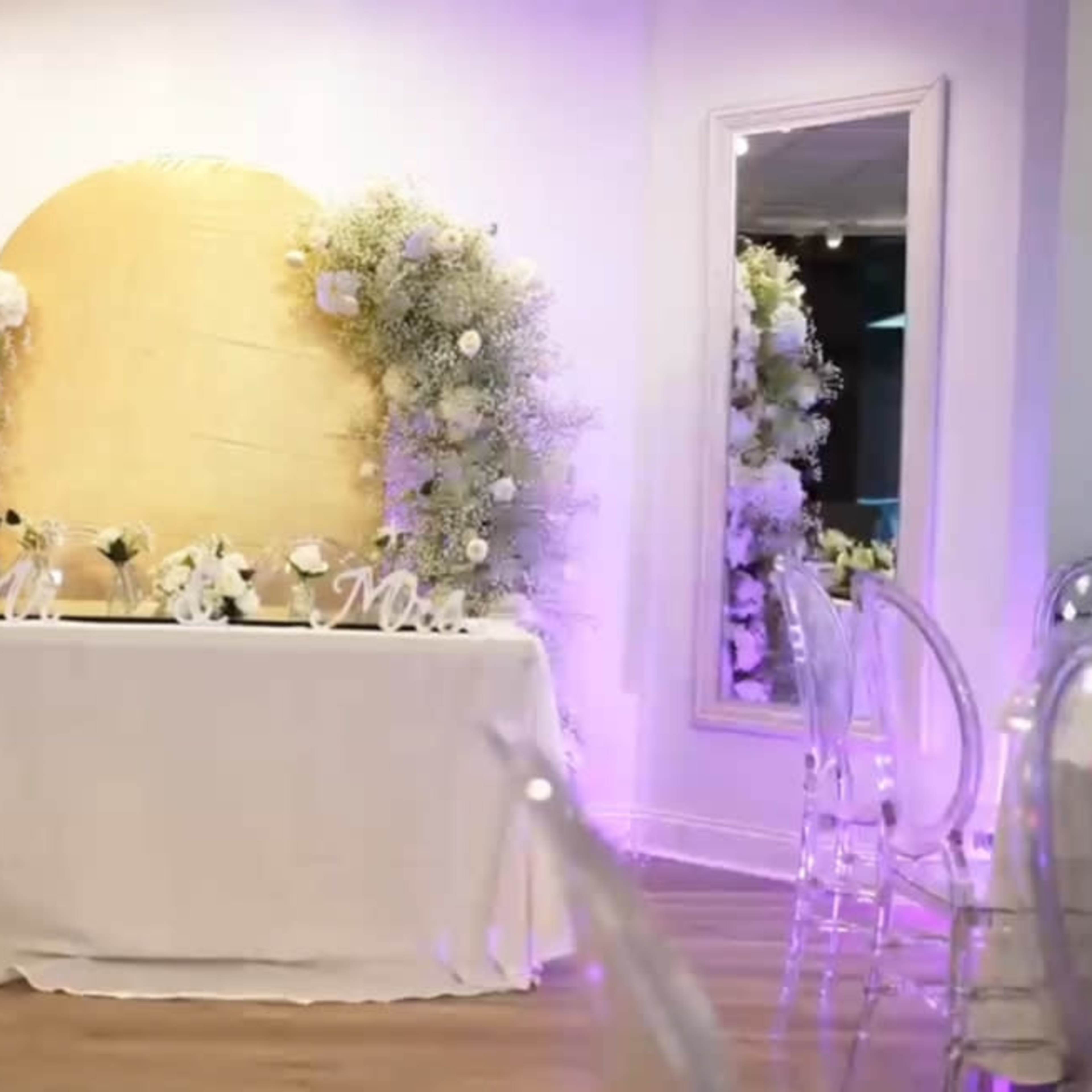 A decorated event space with a golden backdrop, floral arrangements, and a table set with white cloth and decorative items.