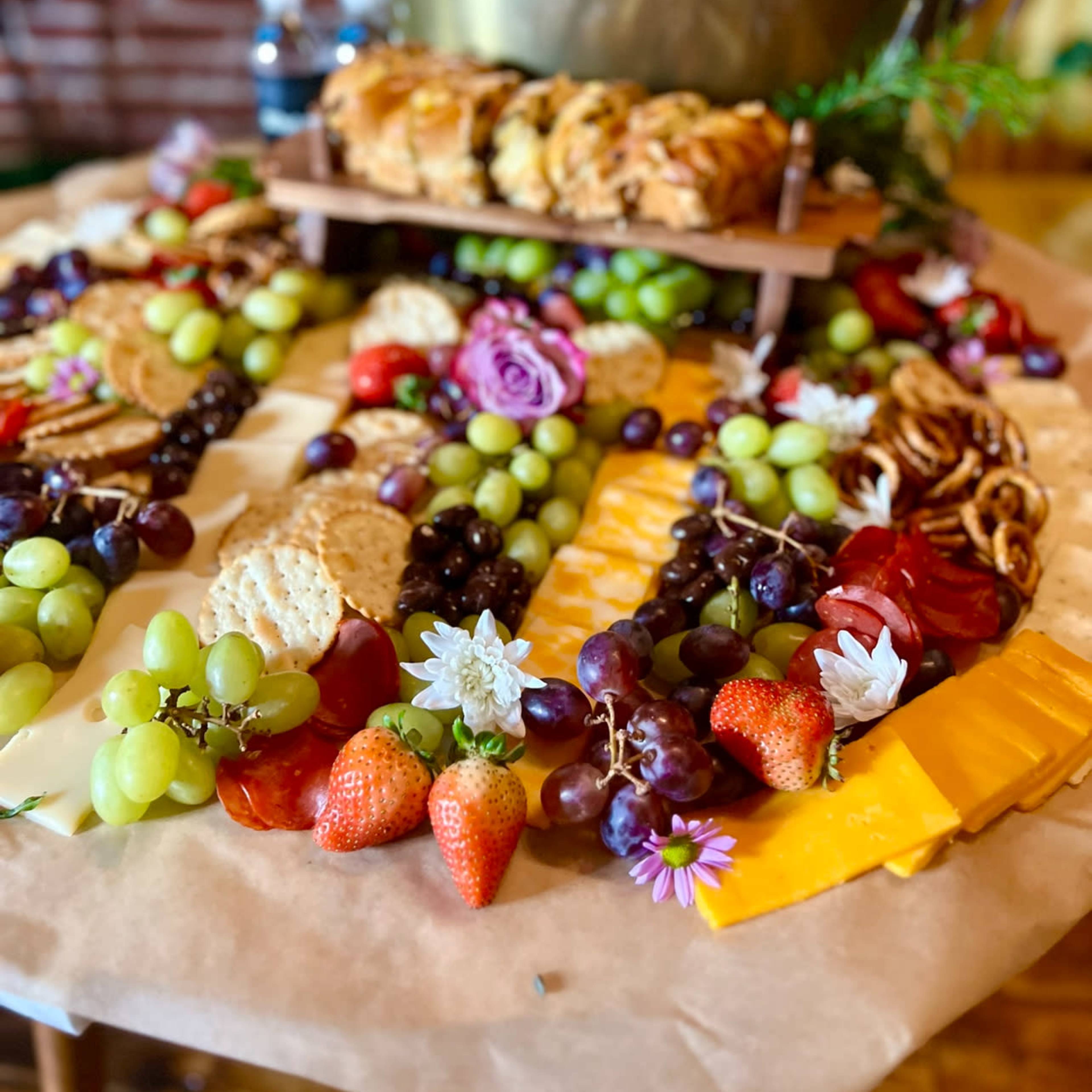 A large, decorative charcuterie board features a variety of fruits, cheeses, crackers, and pastries arranged around a central wooden stand.