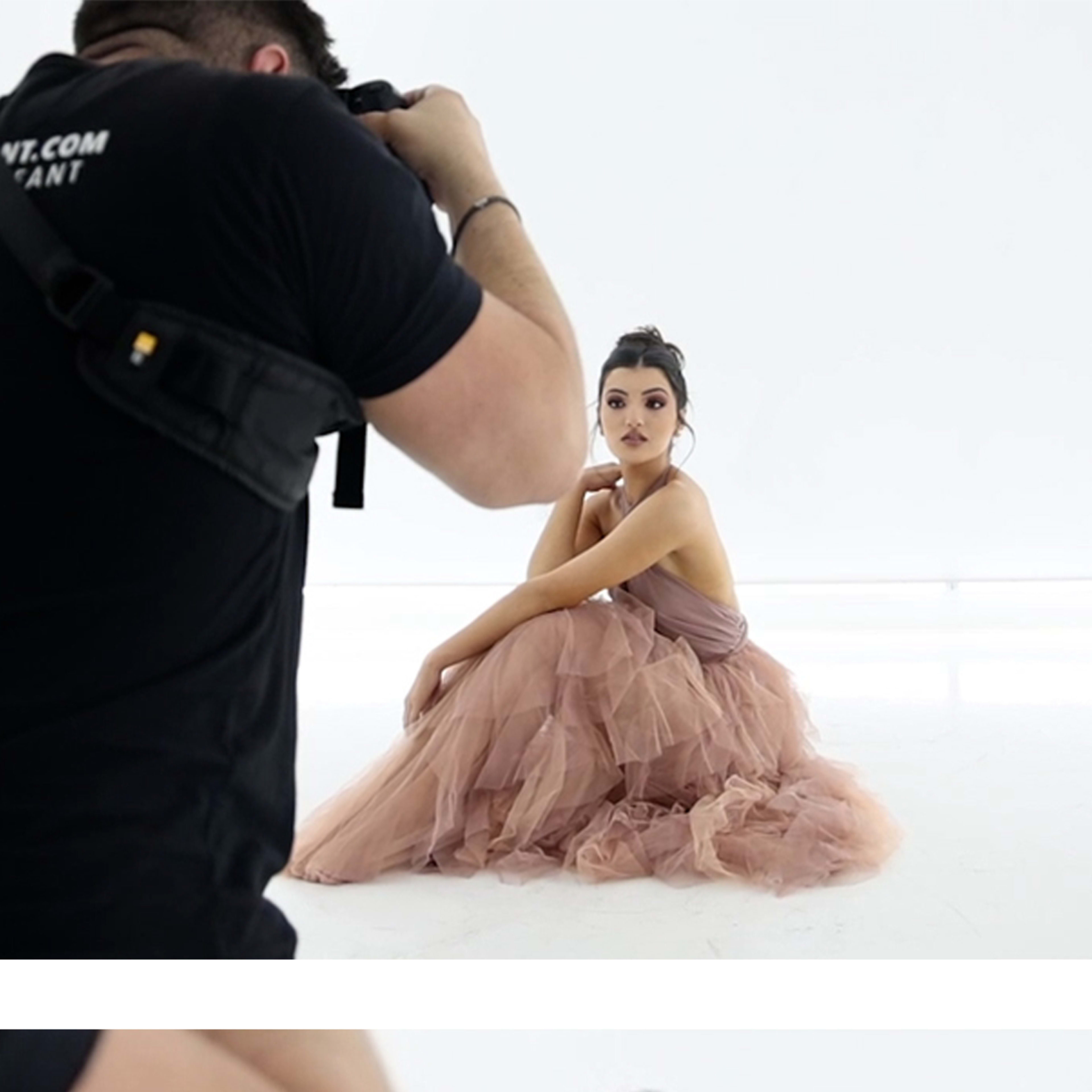A model poses in a flowing, light pink dress while being photographed in a bright, minimalist studio.