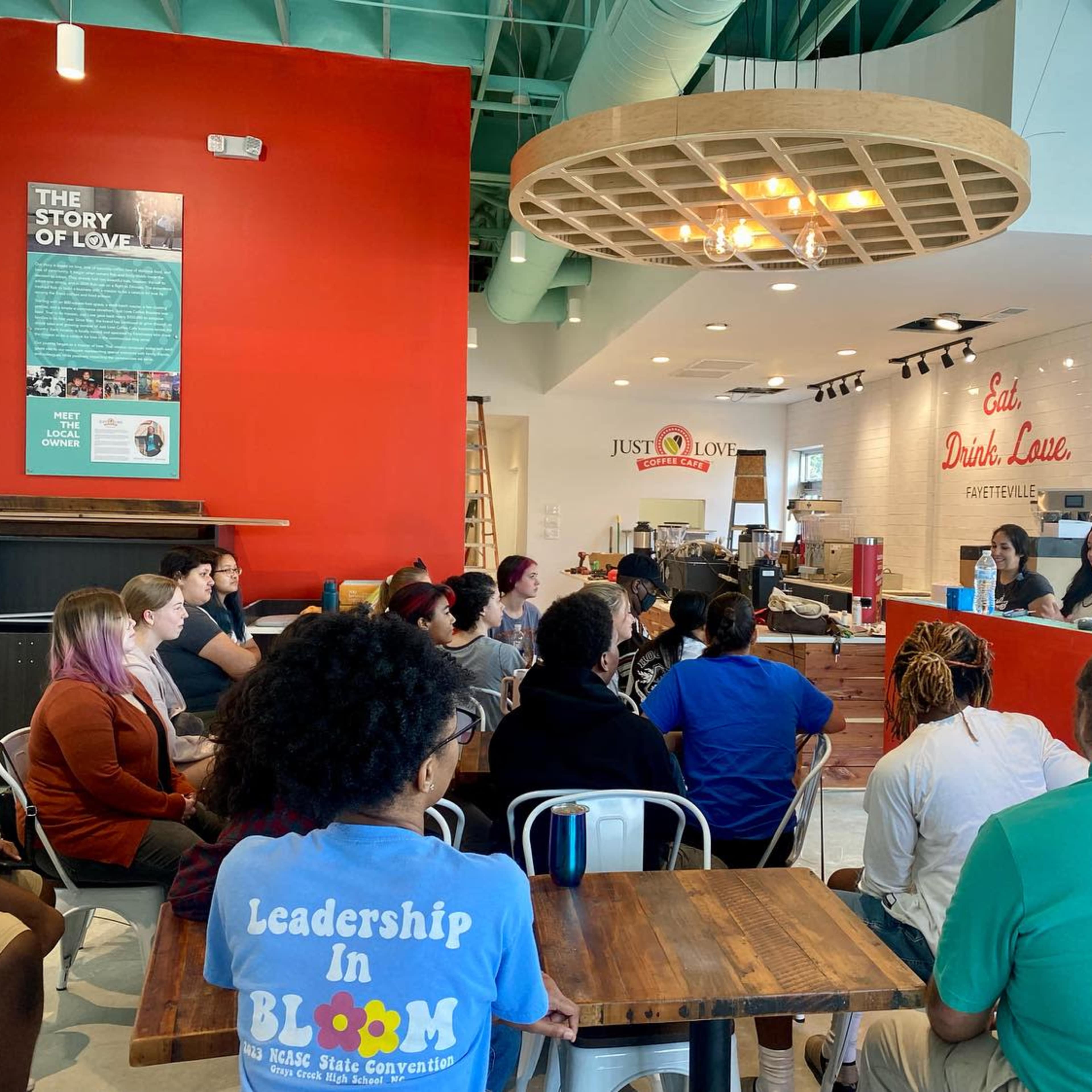A diverse group of people is seated in a cafe, attentively listening to two speakers at the counter.