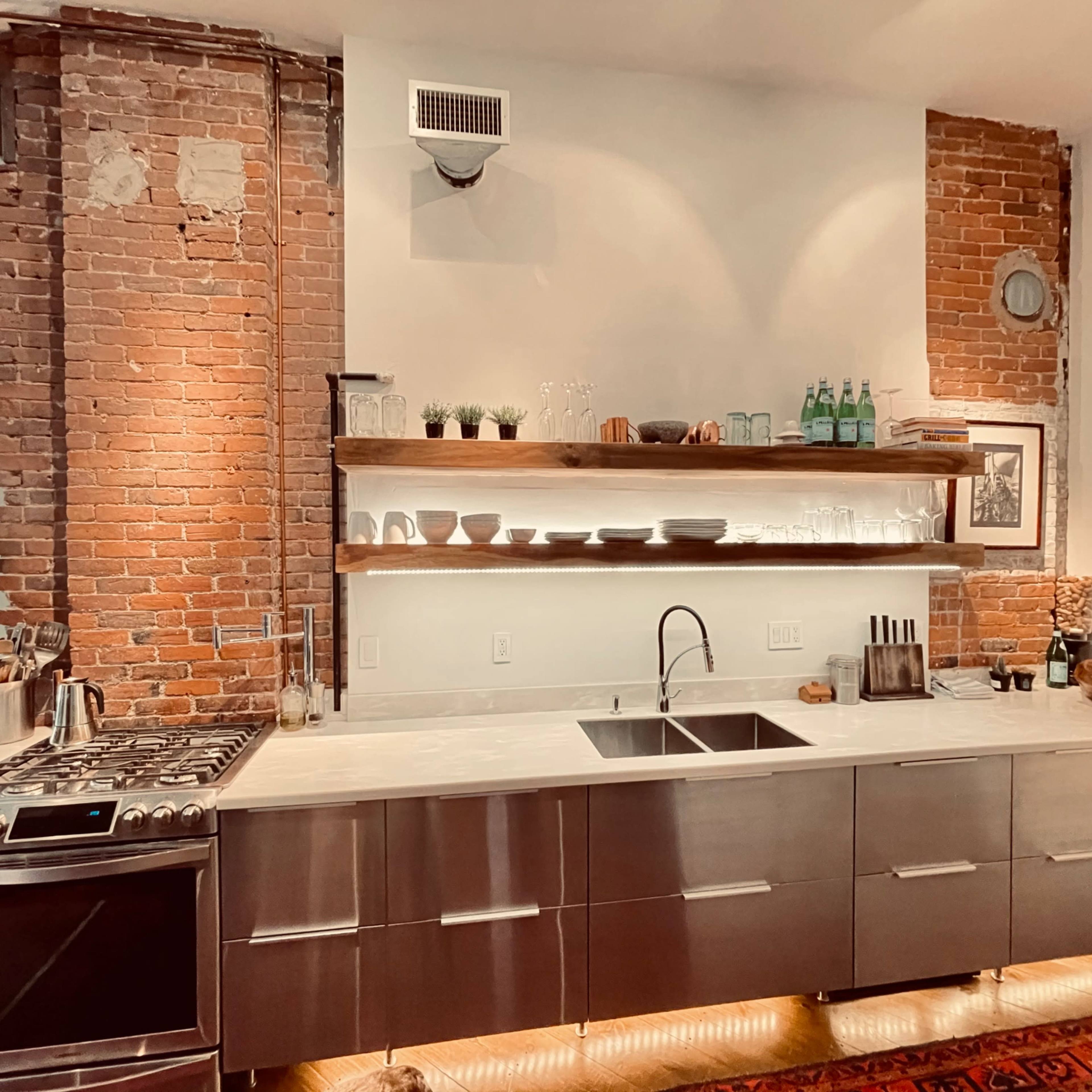 A modern kitchen with exposed brick walls, a stainless steel stove, and a countertop that includes a double sink and open shelving displaying dishes and plants.