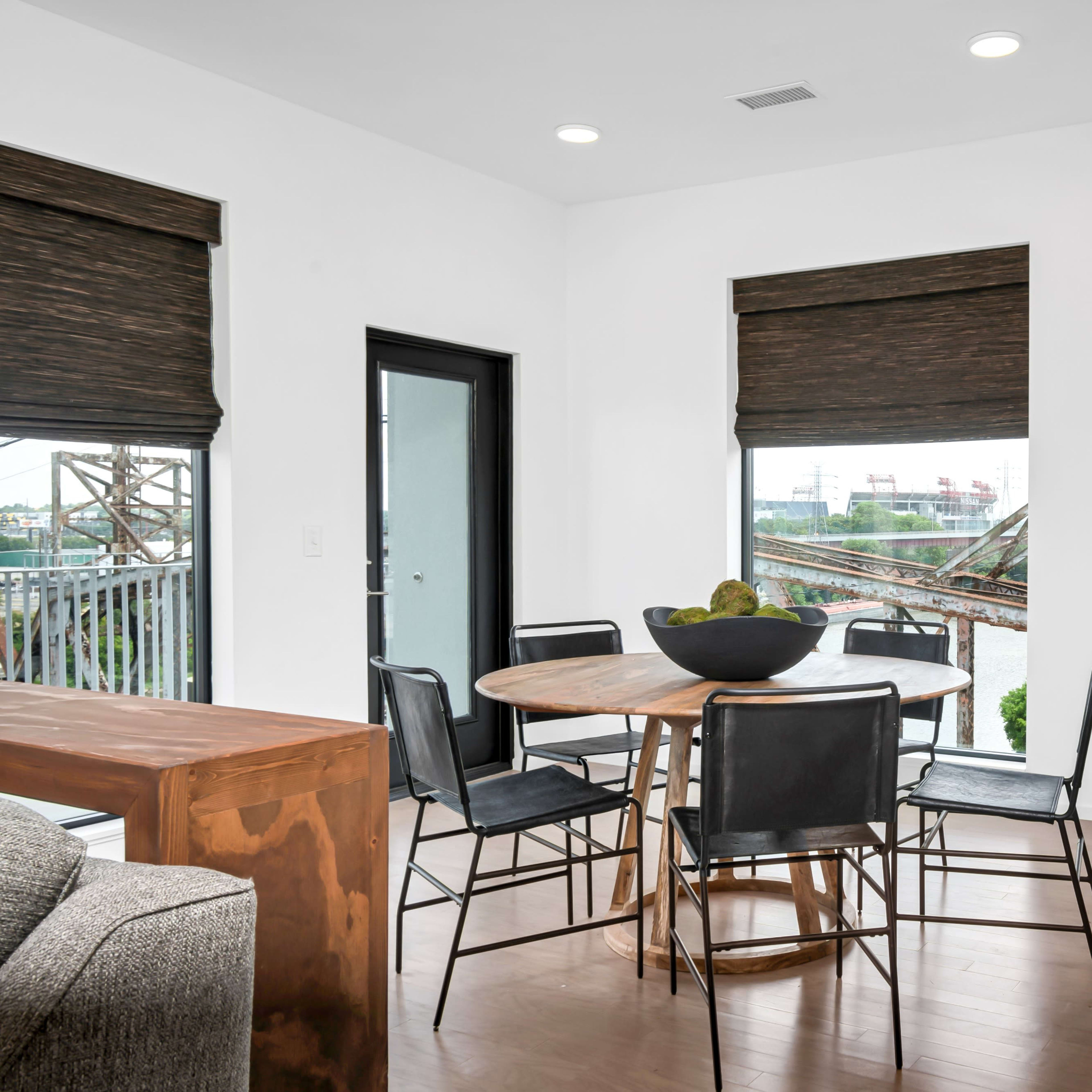 A modern dining area with a round table surrounded by chairs, large windows with shades, and a view of an industrial landscape outside.
