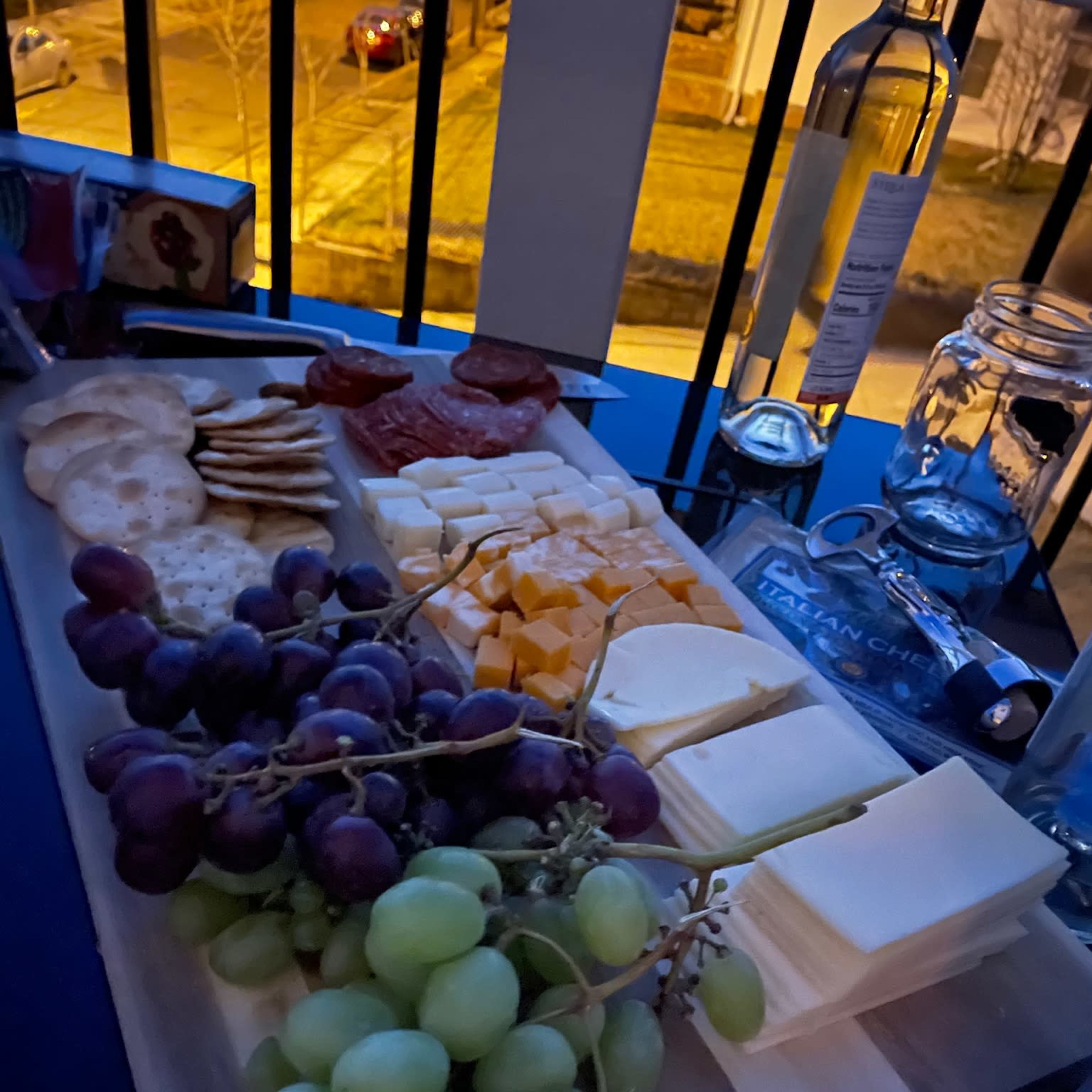 A wooden board displays a variety of cheeses, crackers, sliced meats, and grapes in a dimly lit setting.