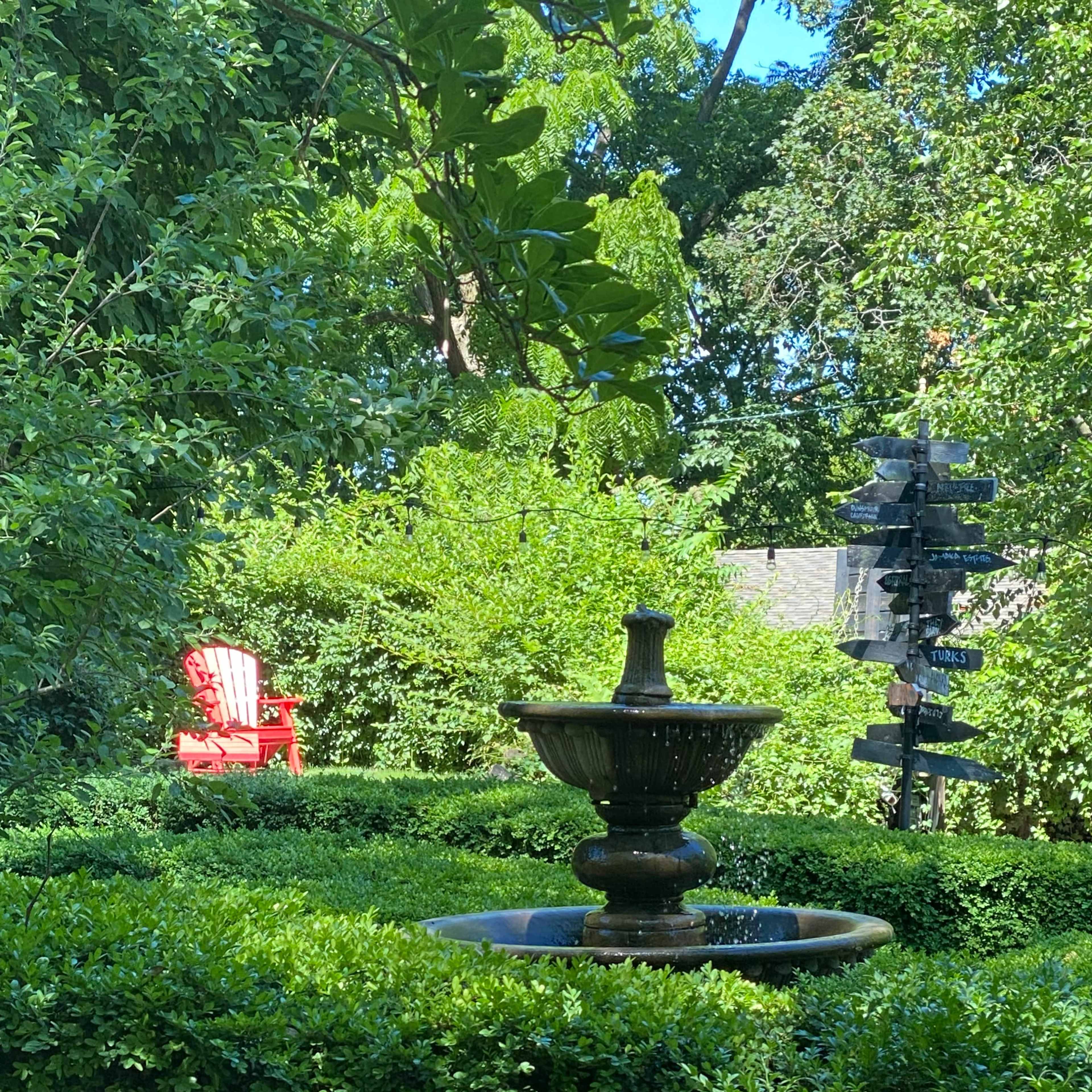 A fountain surrounded by neatly trimmed greenery is in the foreground, with a red chair visible in the background.