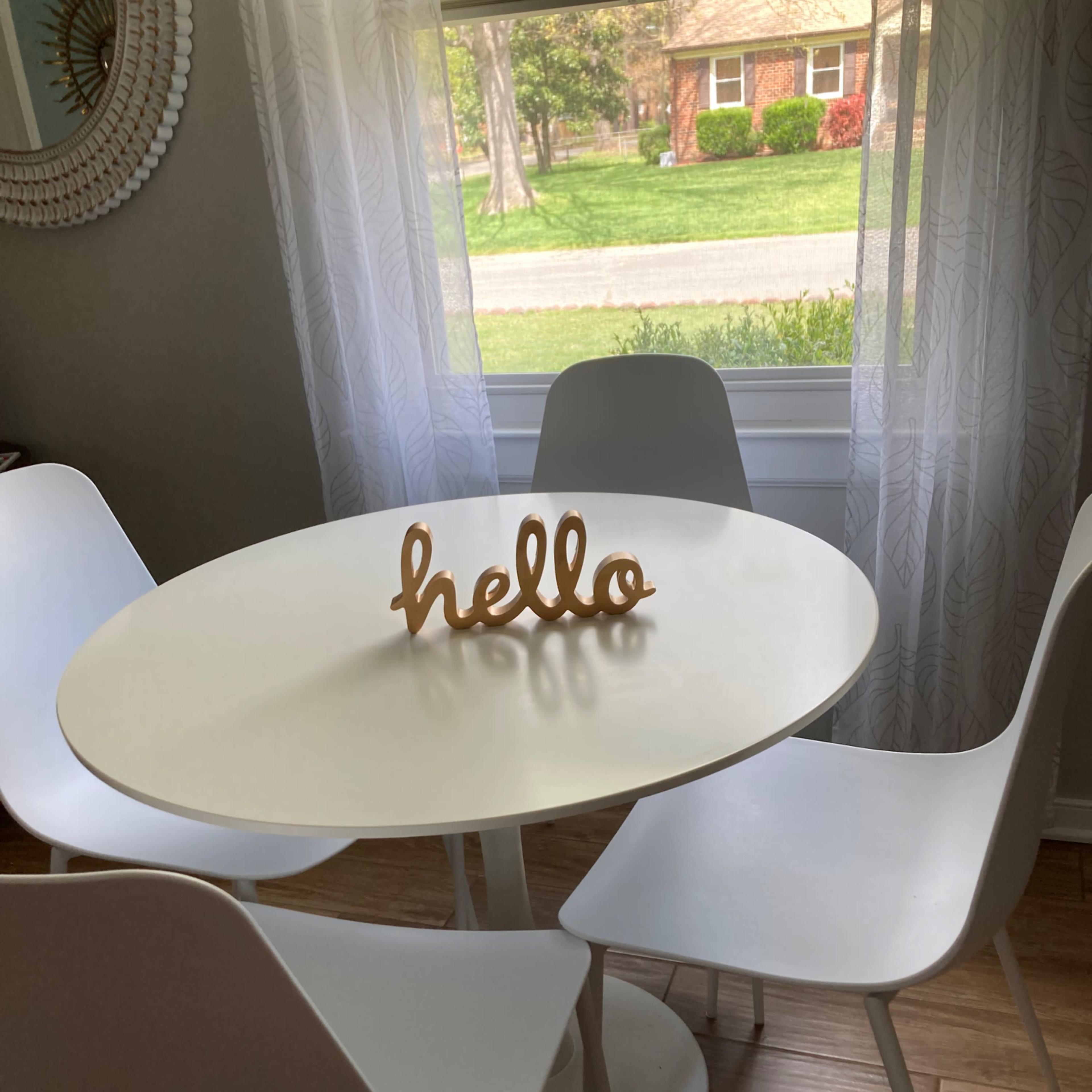 A round white table with four white chairs and a decorative "hello" greeting sits in front of a window with sheer curtains.