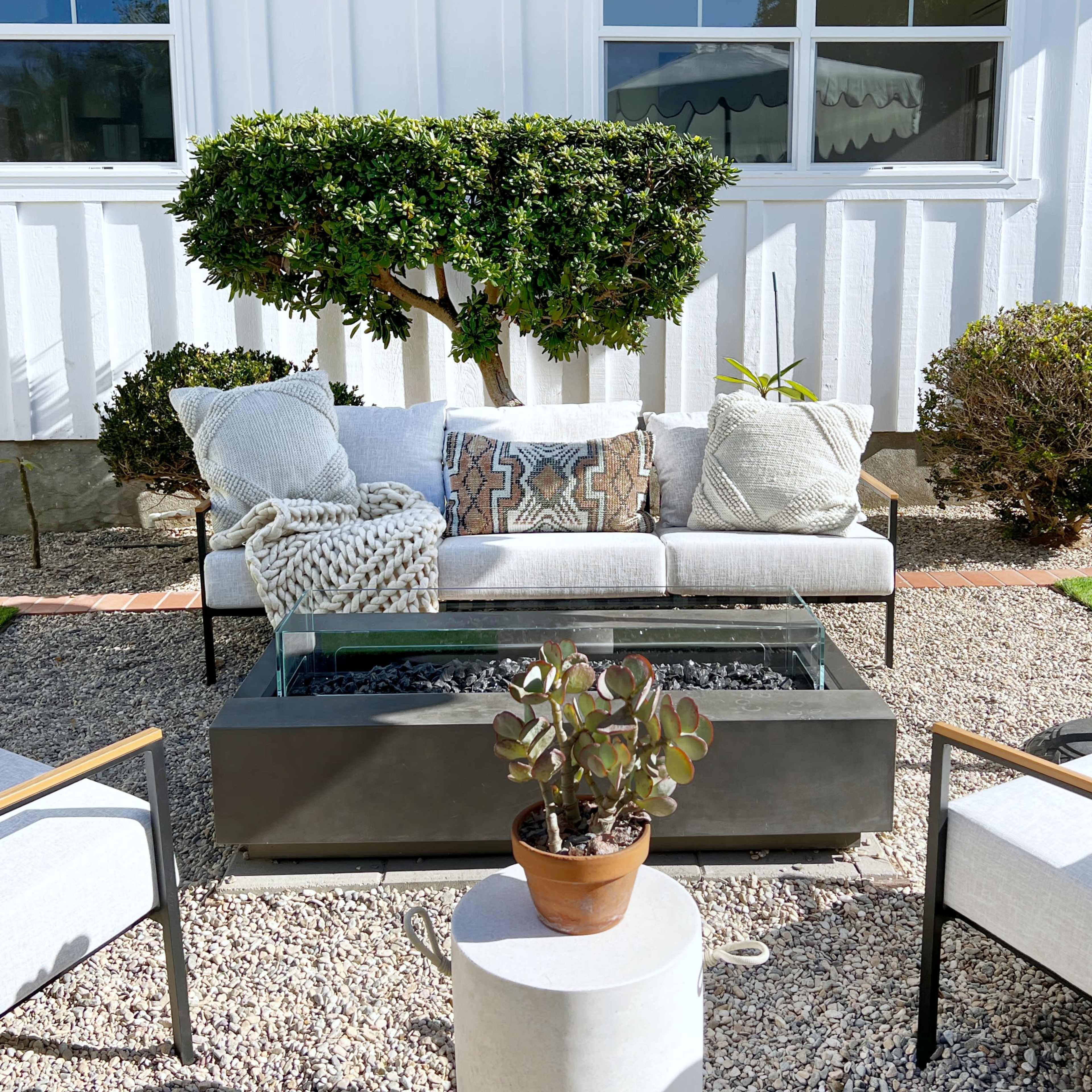 A modern outdoor seating area featuring a sofa with decorative pillows, a coffee table with a glass top, and potted plants in a gravel landscape.