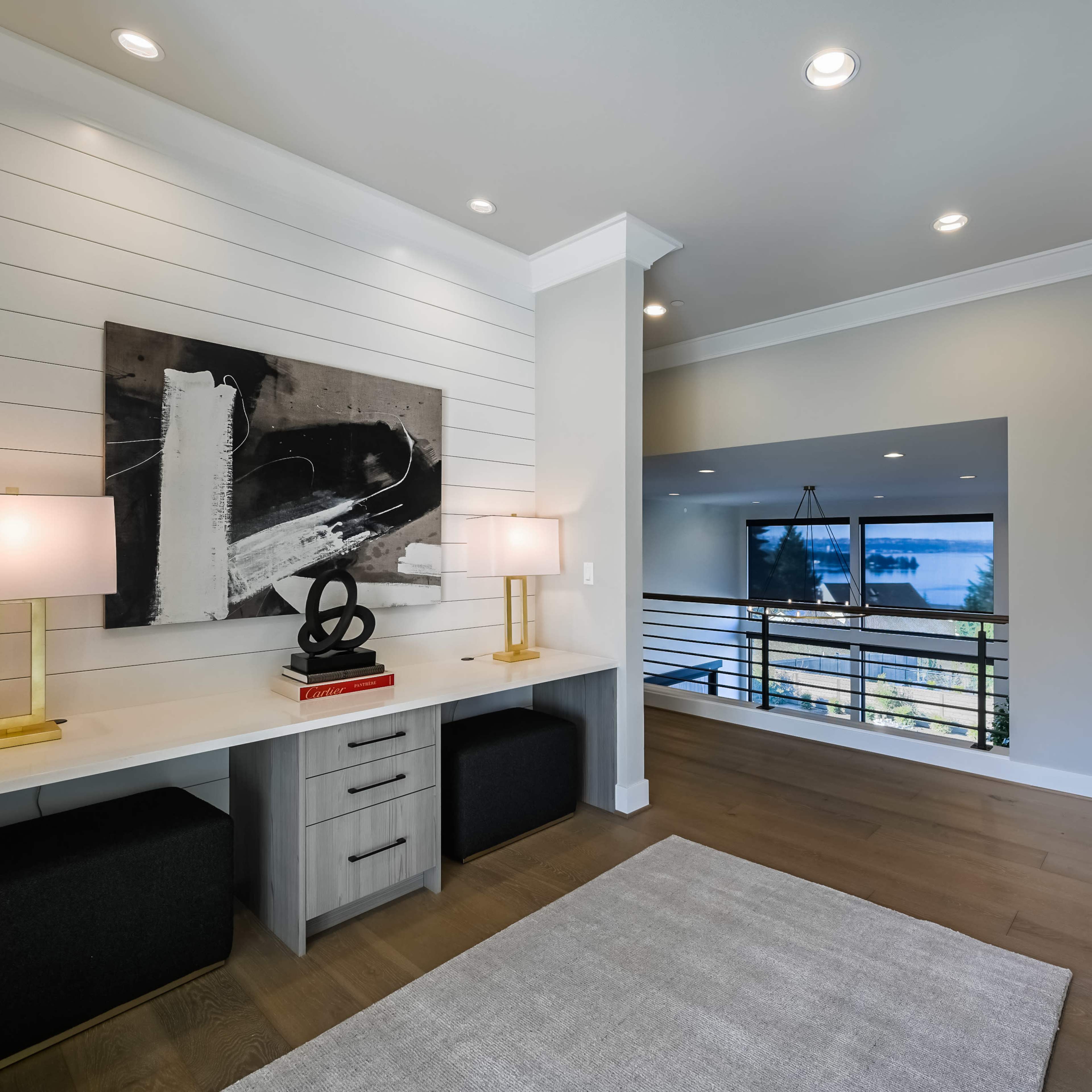 A modern interior space featuring a minimalistic desk with lamps, artwork on the wall, and a view of a lake through a large window.