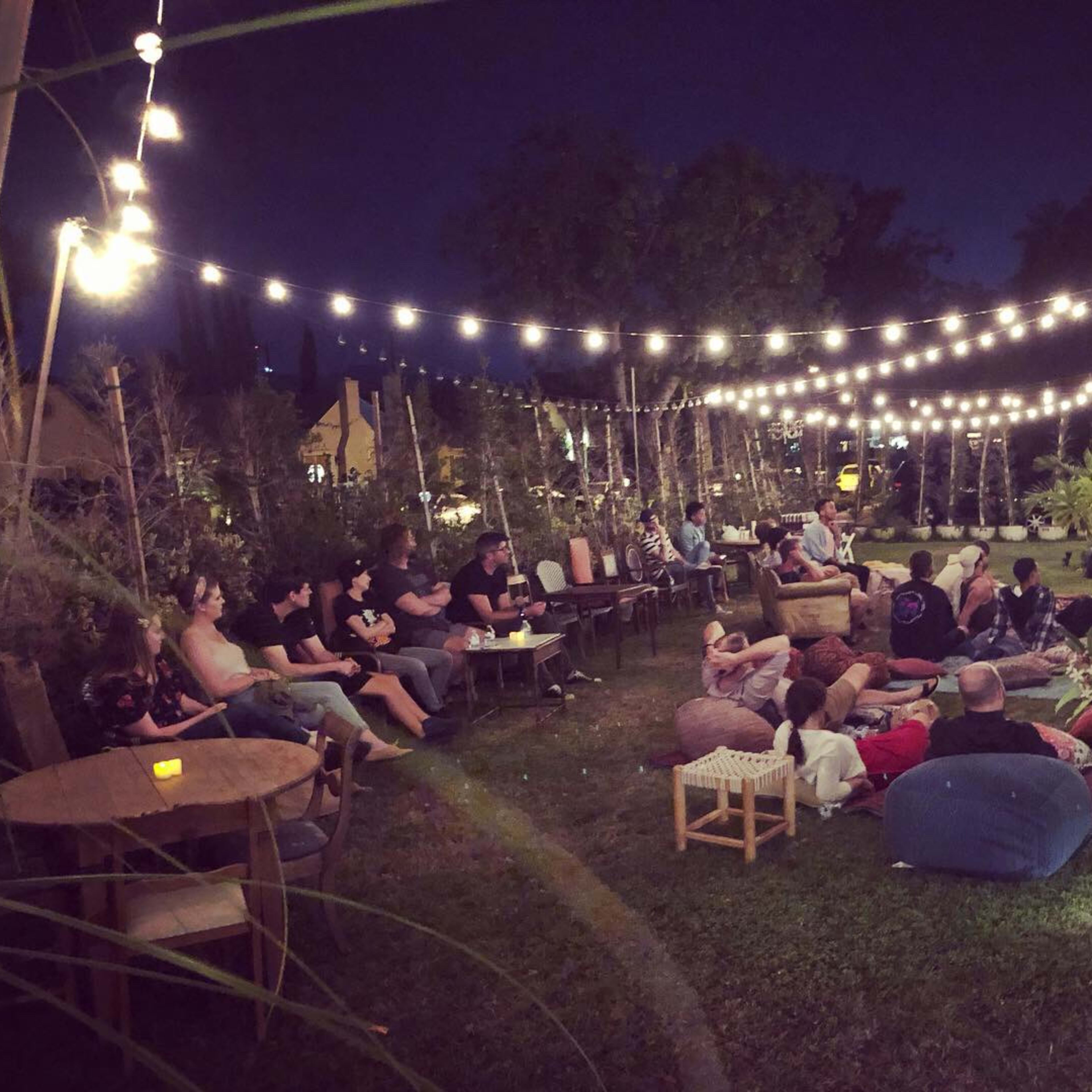 A group of people sits in a garden under string lights, gathered for an outdoor performance.