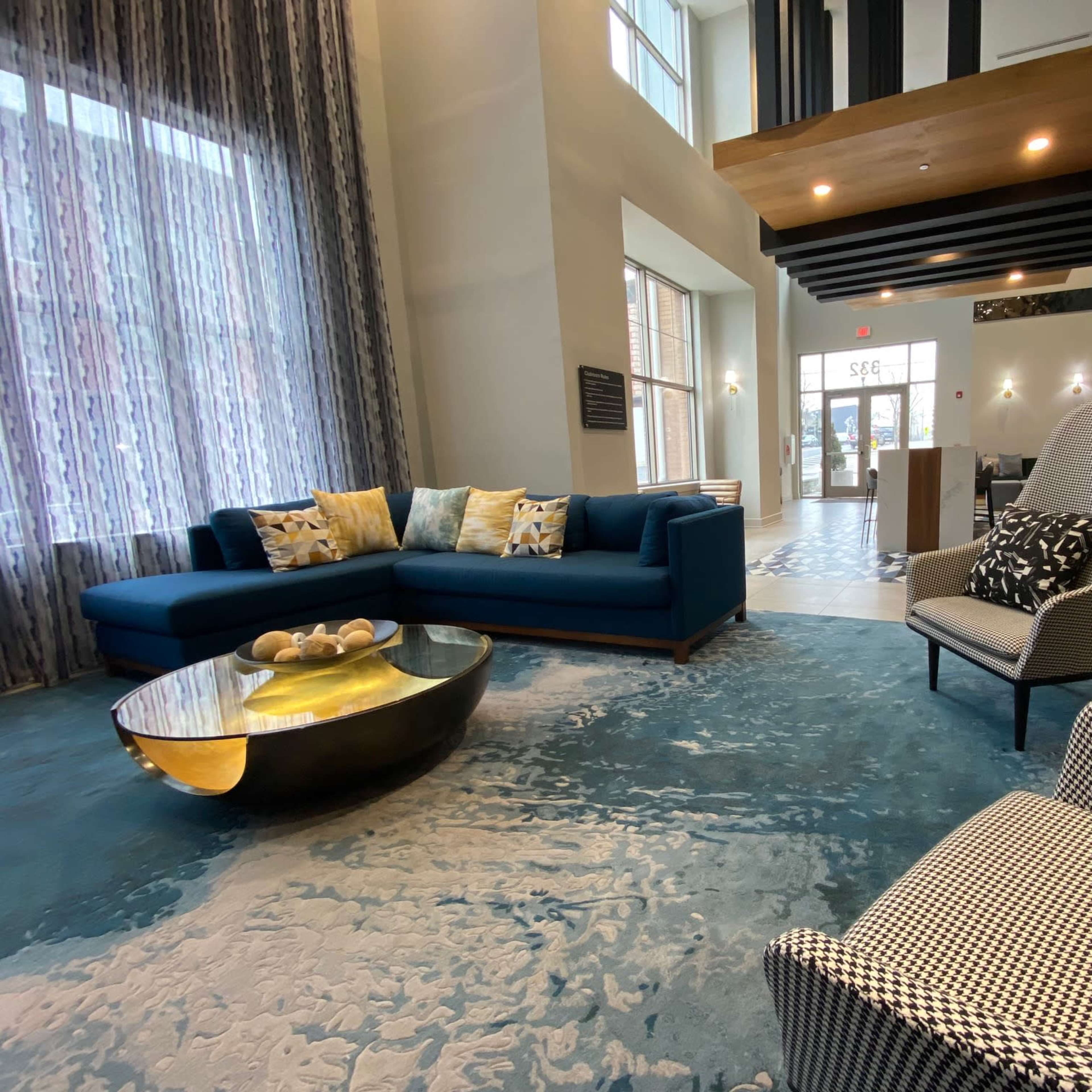 A modern lobby area with a blue sectional sofa, a round coffee table, and large windows allowing natural light to fill the space.