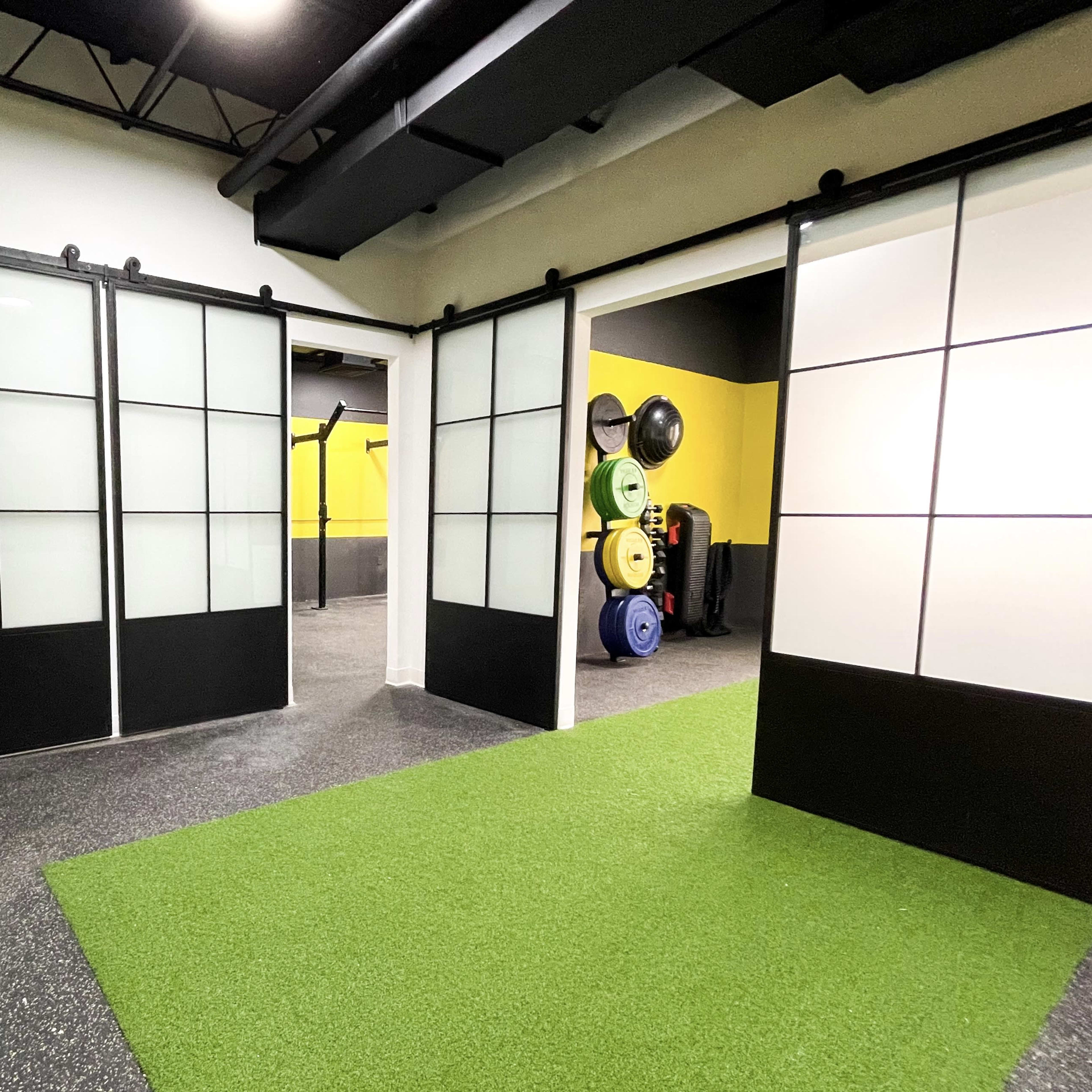 A modern gym interior featuring sliding glass doors, a green turf area, and workout equipment along the walls.