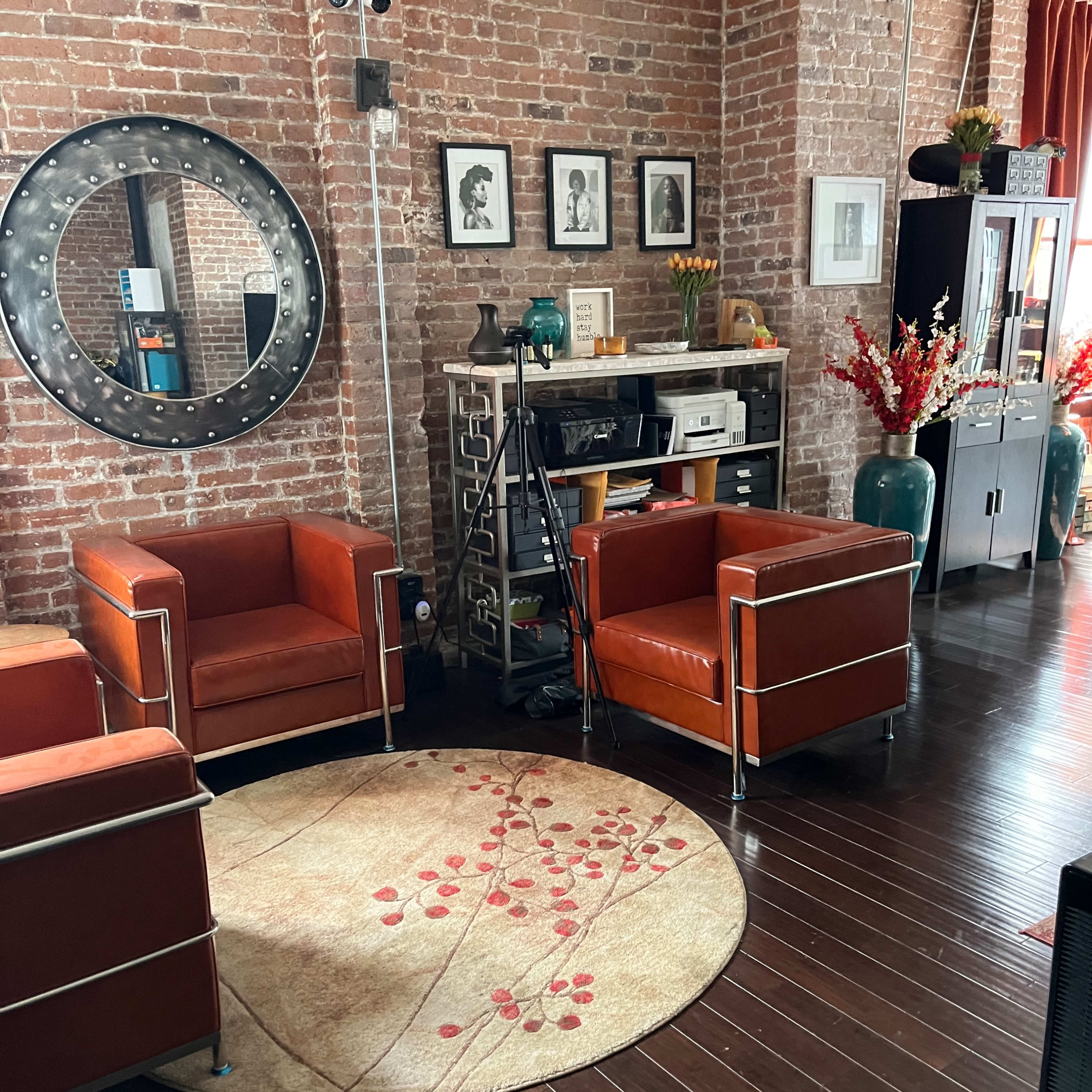 A living room with exposed brick walls, two orange chairs, a round rug with a floral pattern, and a wooden side table displaying framed artwork.