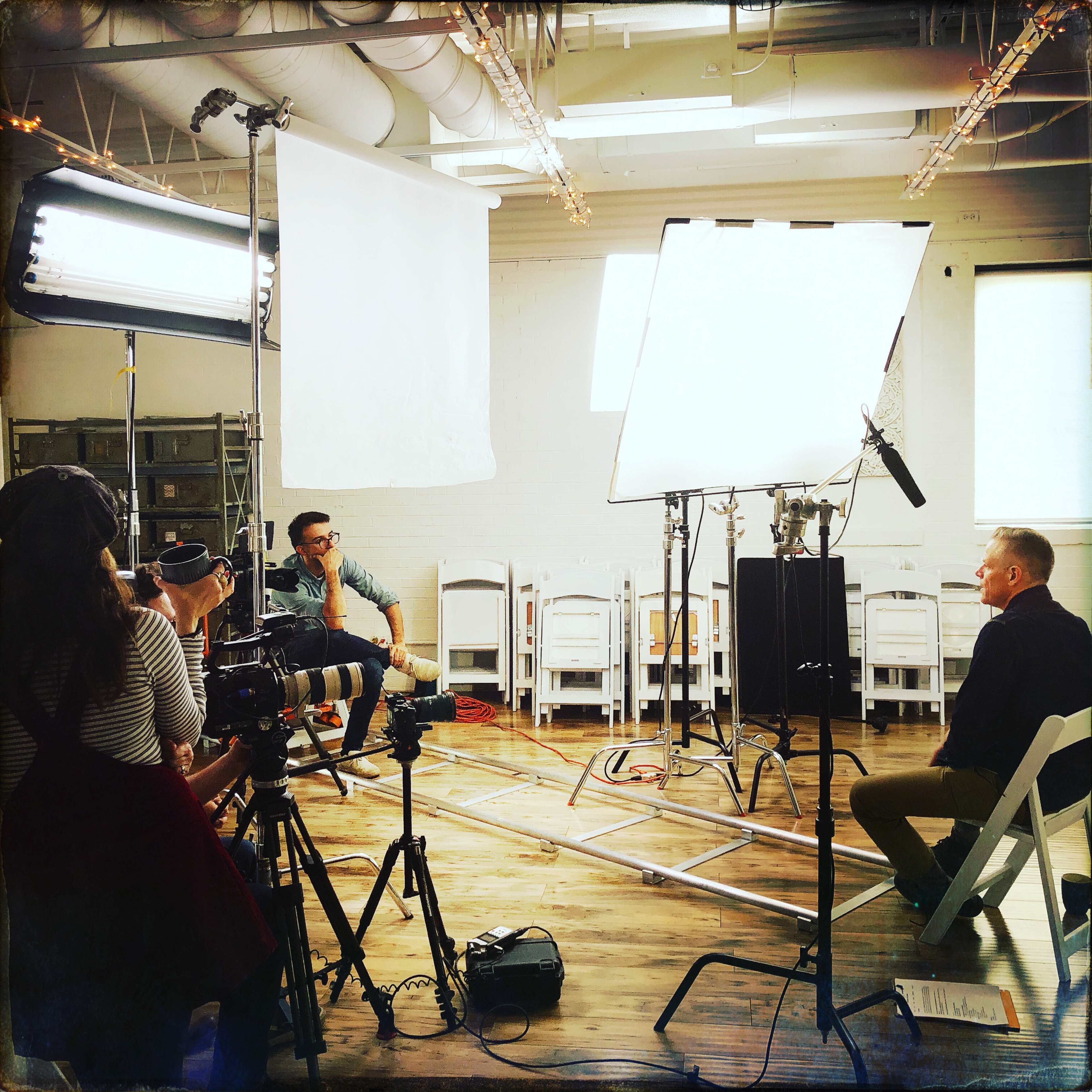 A film crew is set up in a bright room to interview a seated subject while another crew member operates a camera.