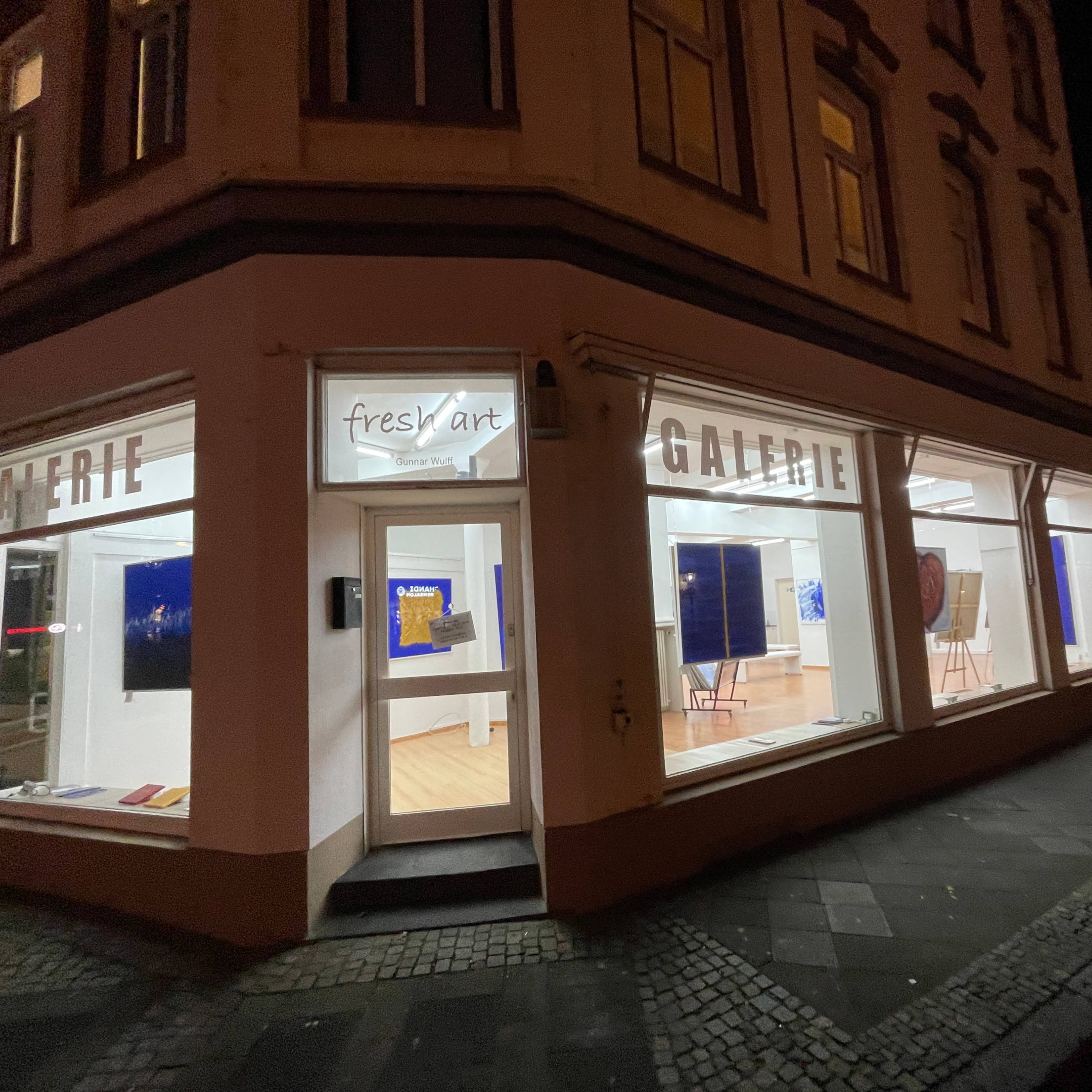 A lit art gallery at night, featuring large windows displaying various artworks.