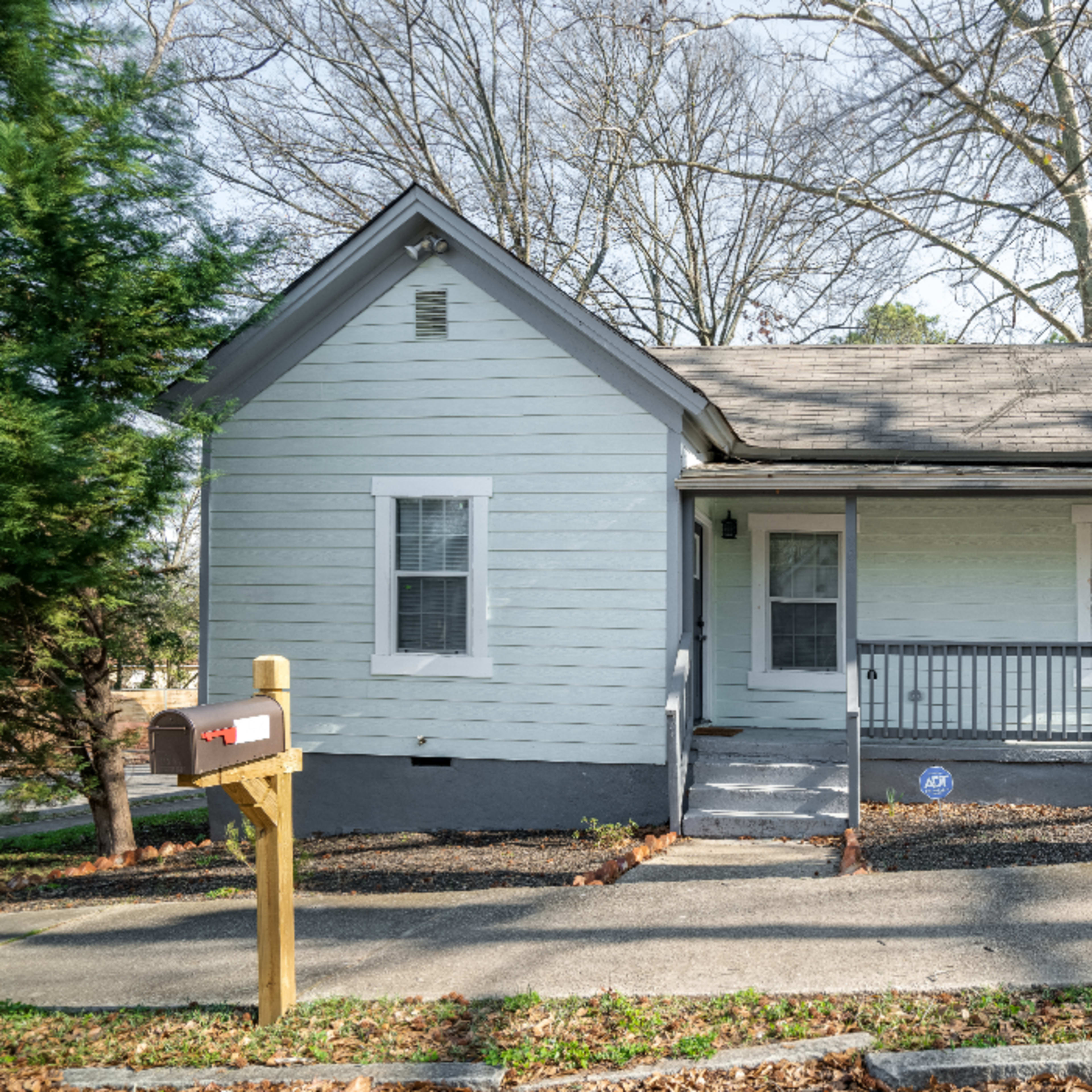 A small pale blue house with a front porch and a gravel driveway is situated next to a large tree.