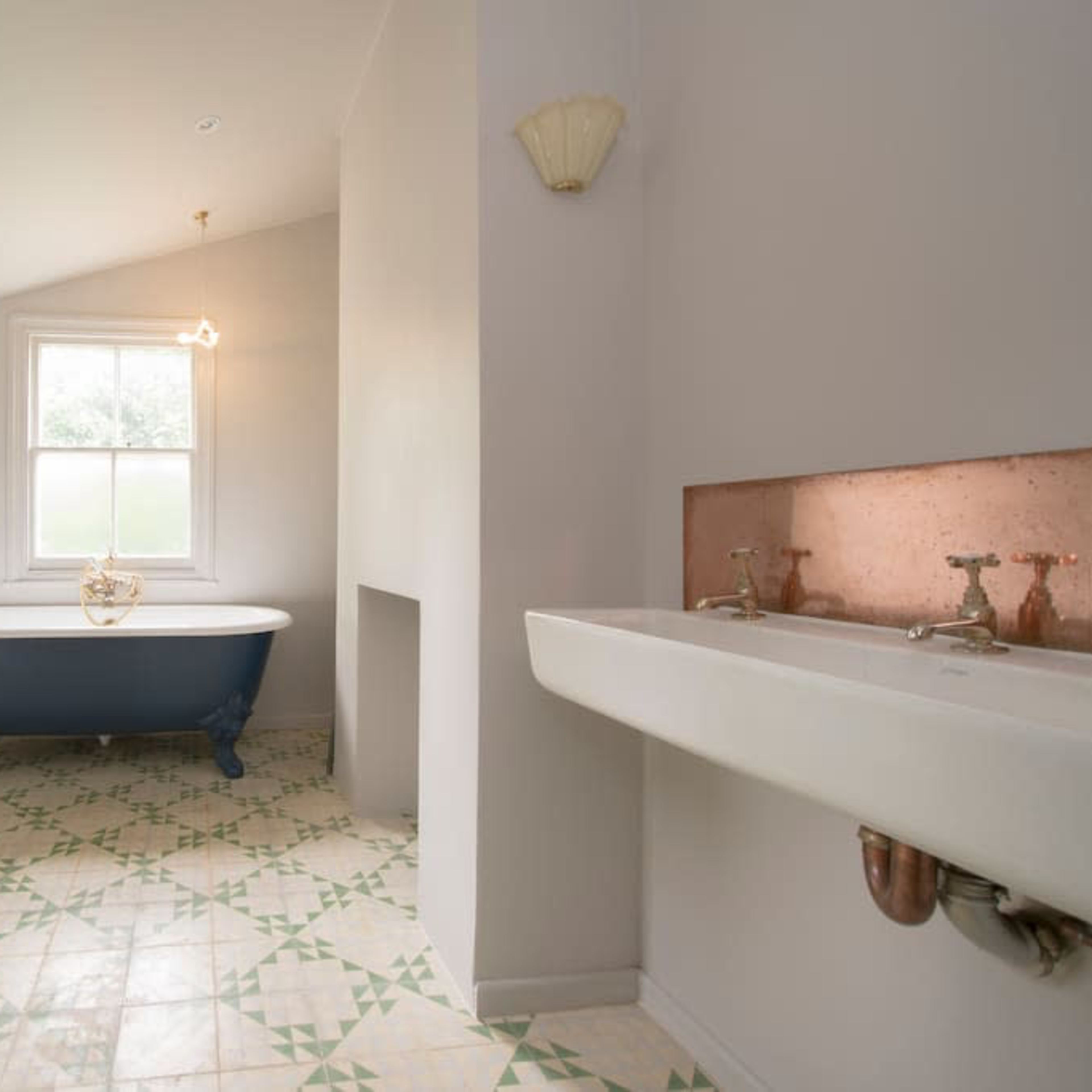 A bathroom featuring a blue freestanding bathtub, a white toilet, and a long sink with bronze faucets, all set against a light gray wall and patterned tile floor.