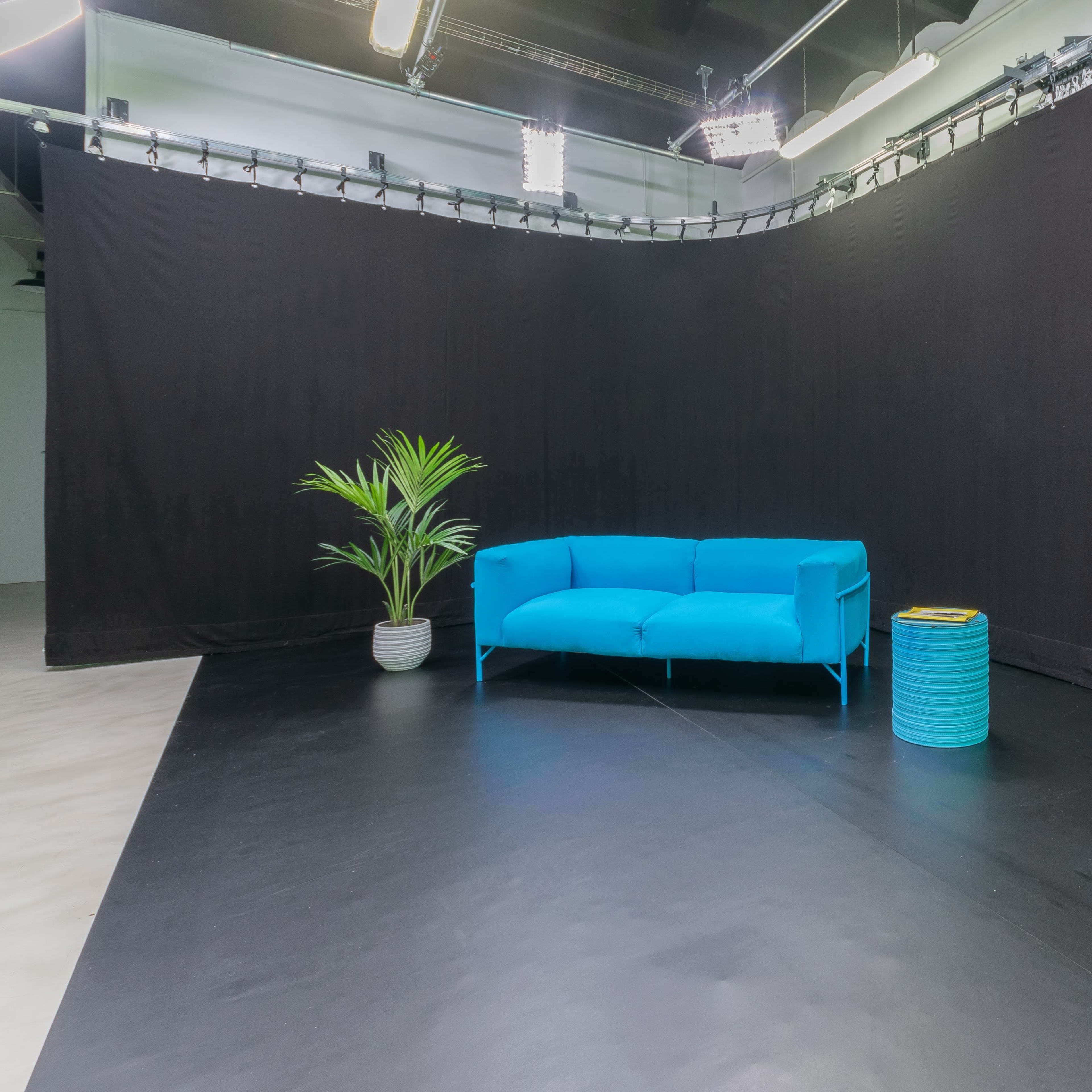 A bright blue sofa and a teal accent table are positioned in front of a black backdrop in a spacious studio setting.
