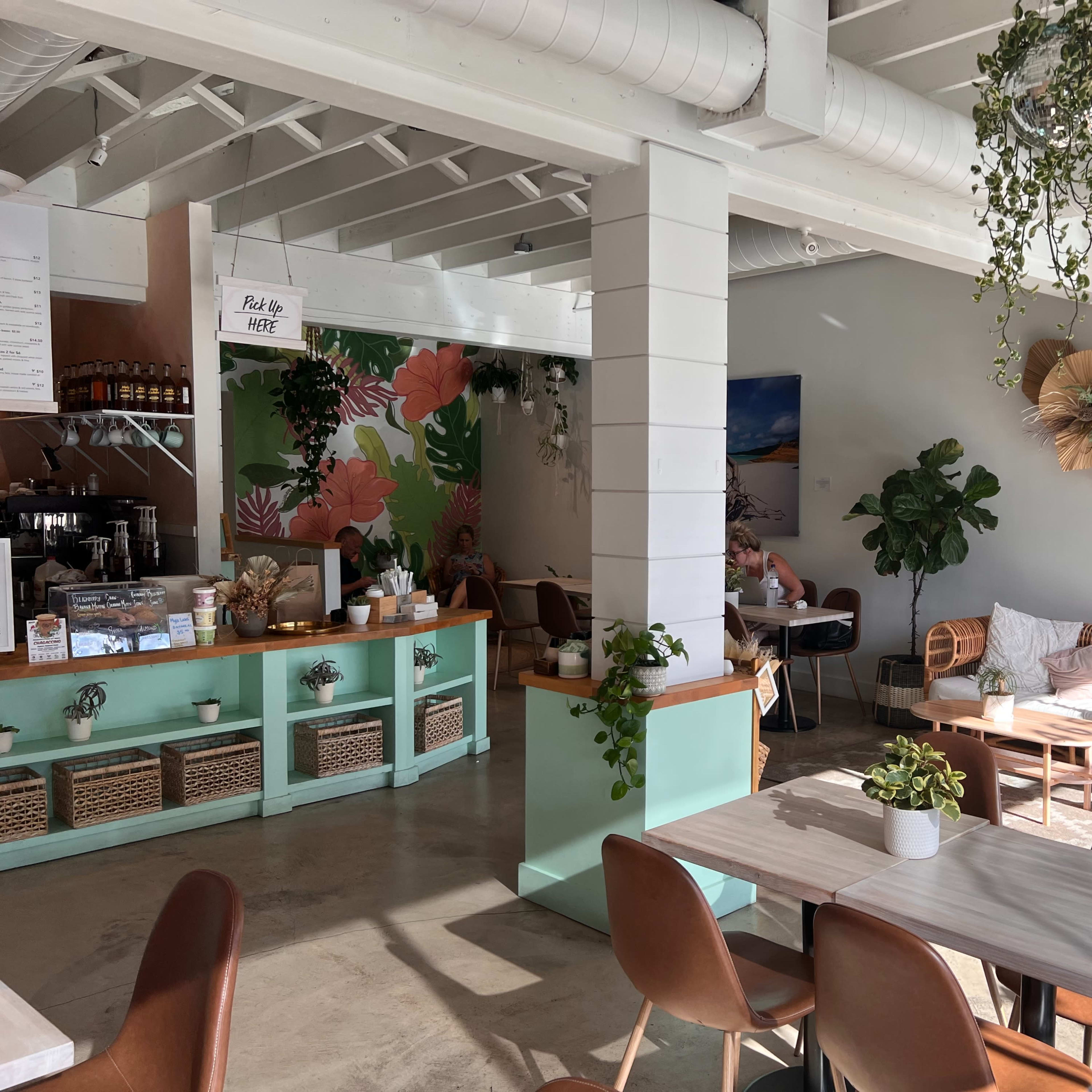 A bright café interior with green accents, plants, and a mix of seating options, including chairs and a cozy lounge area.