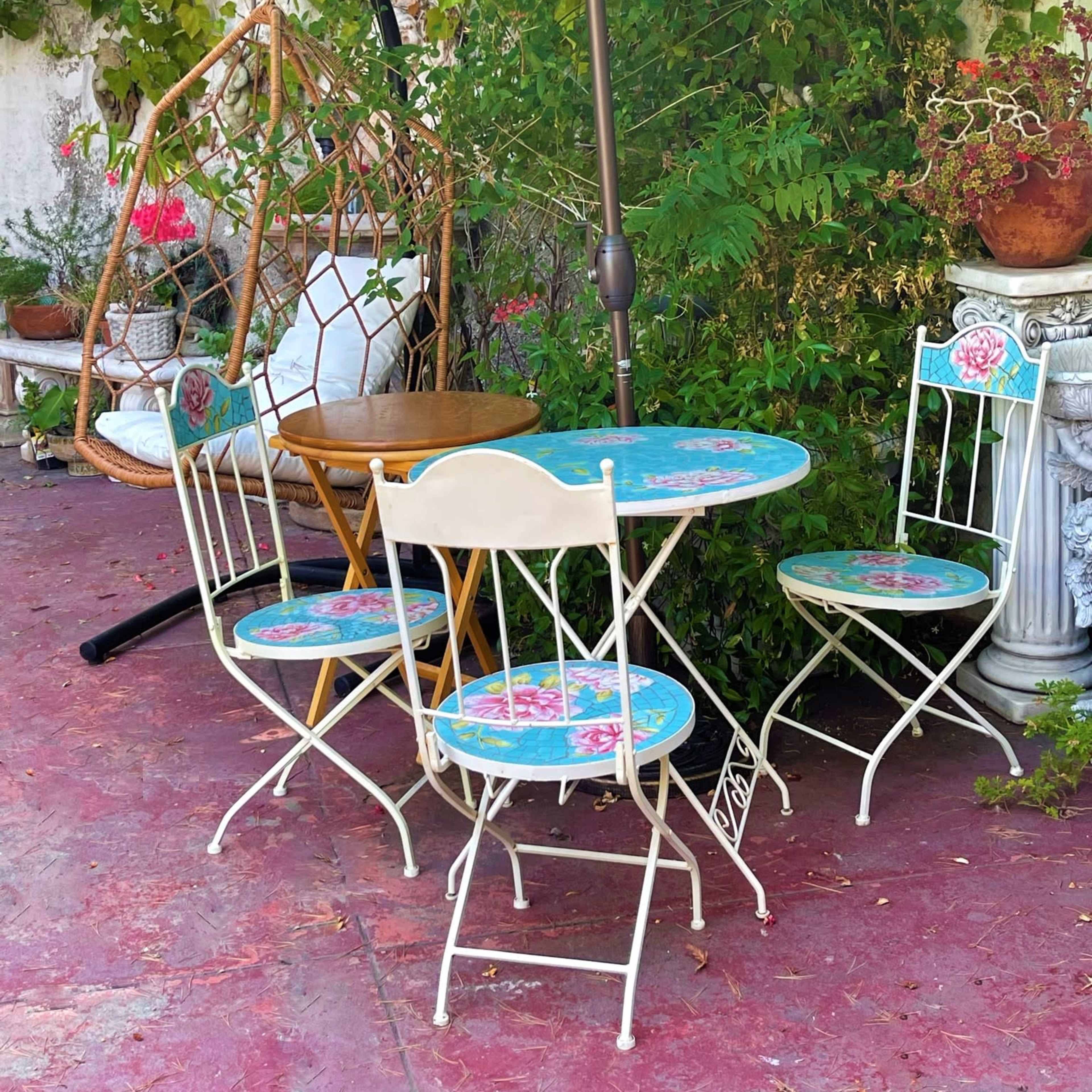 A small outdoor seating area features a table and four chairs under a turquoise umbrella, surrounded by greenery and potted plants.