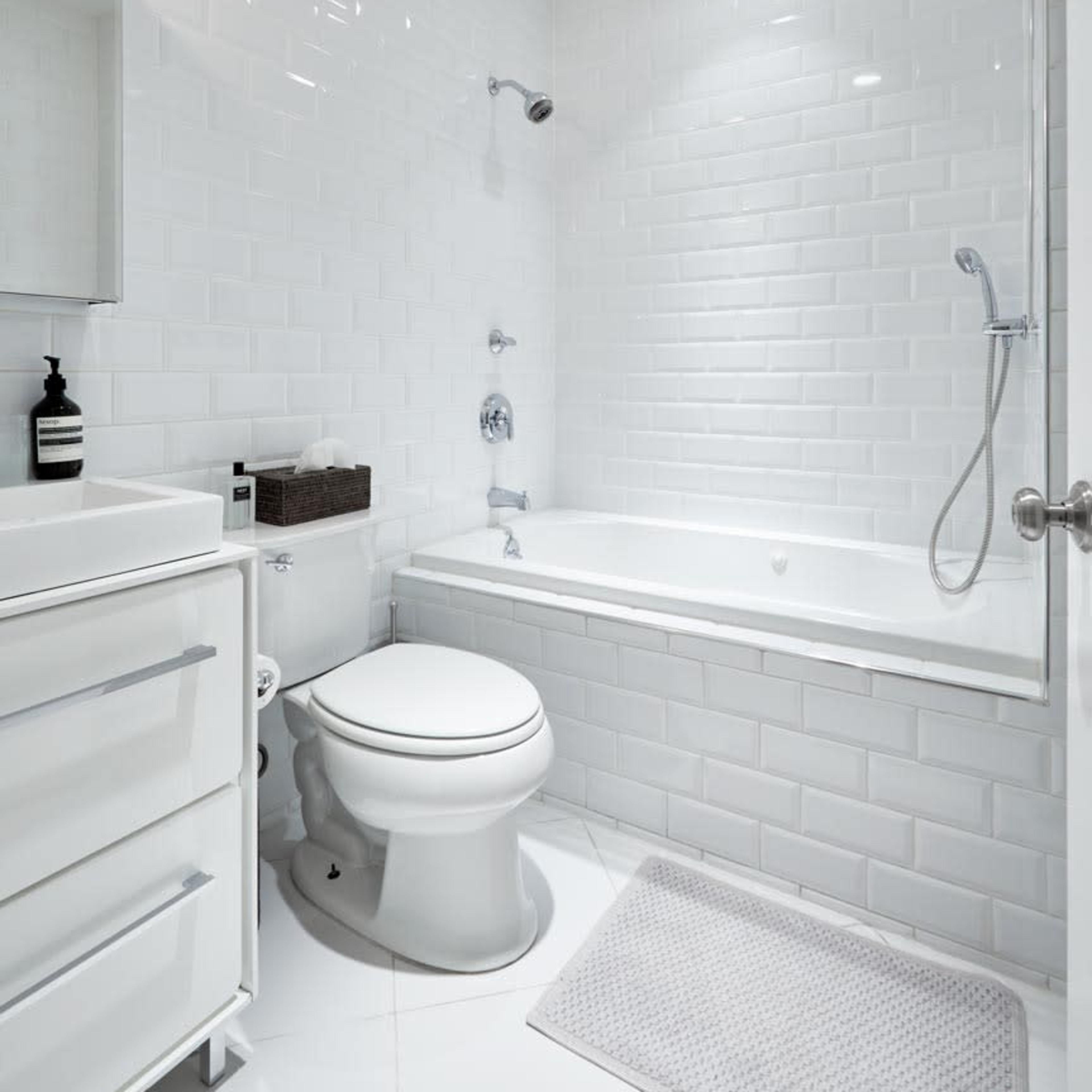 A clean, modern bathroom featuring a bathtub, a toilet, and a sink with storage drawers, all set against white tiled walls.