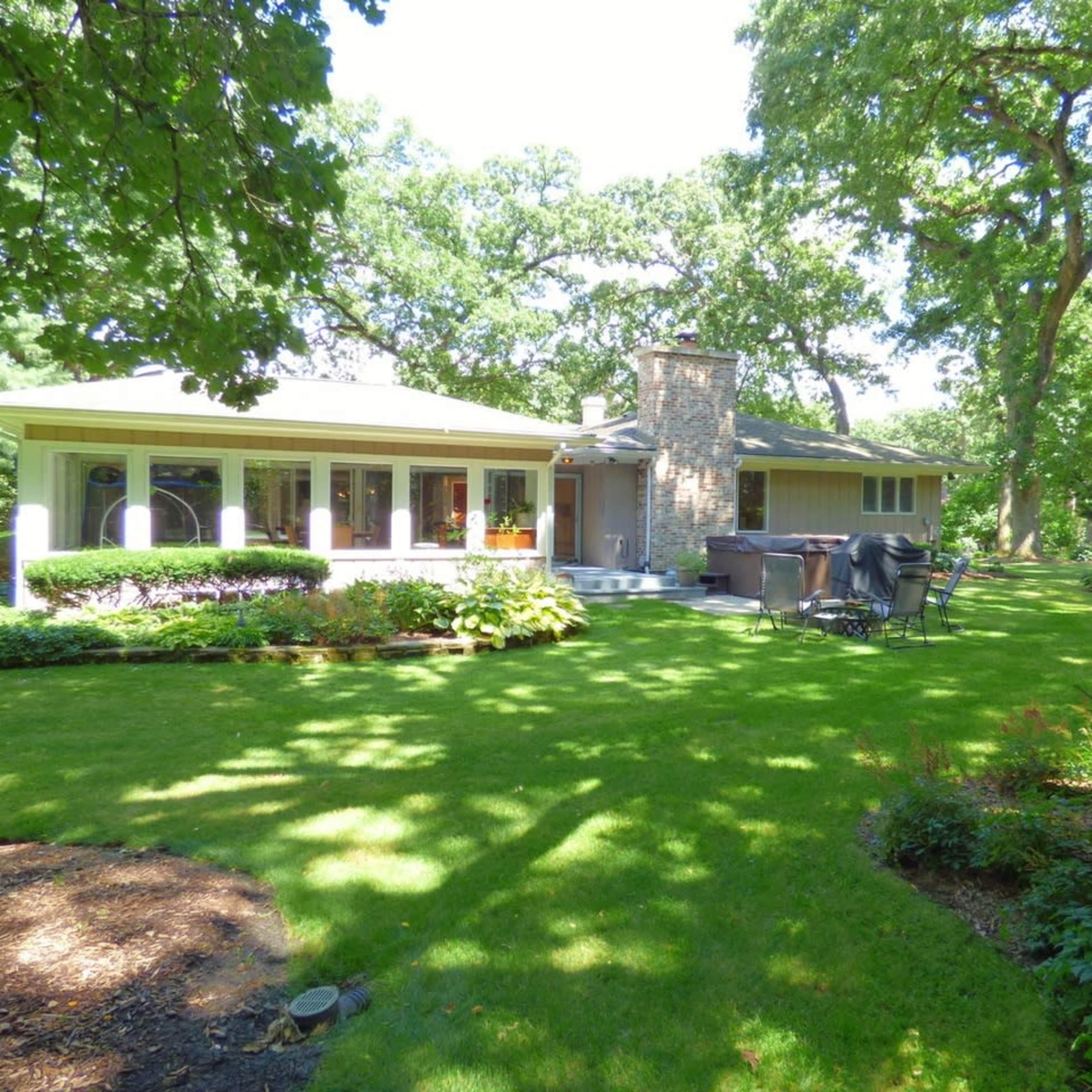 A spacious home with a stone chimney is surrounded by a well-maintained lawn and a variety of trees and shrubs.