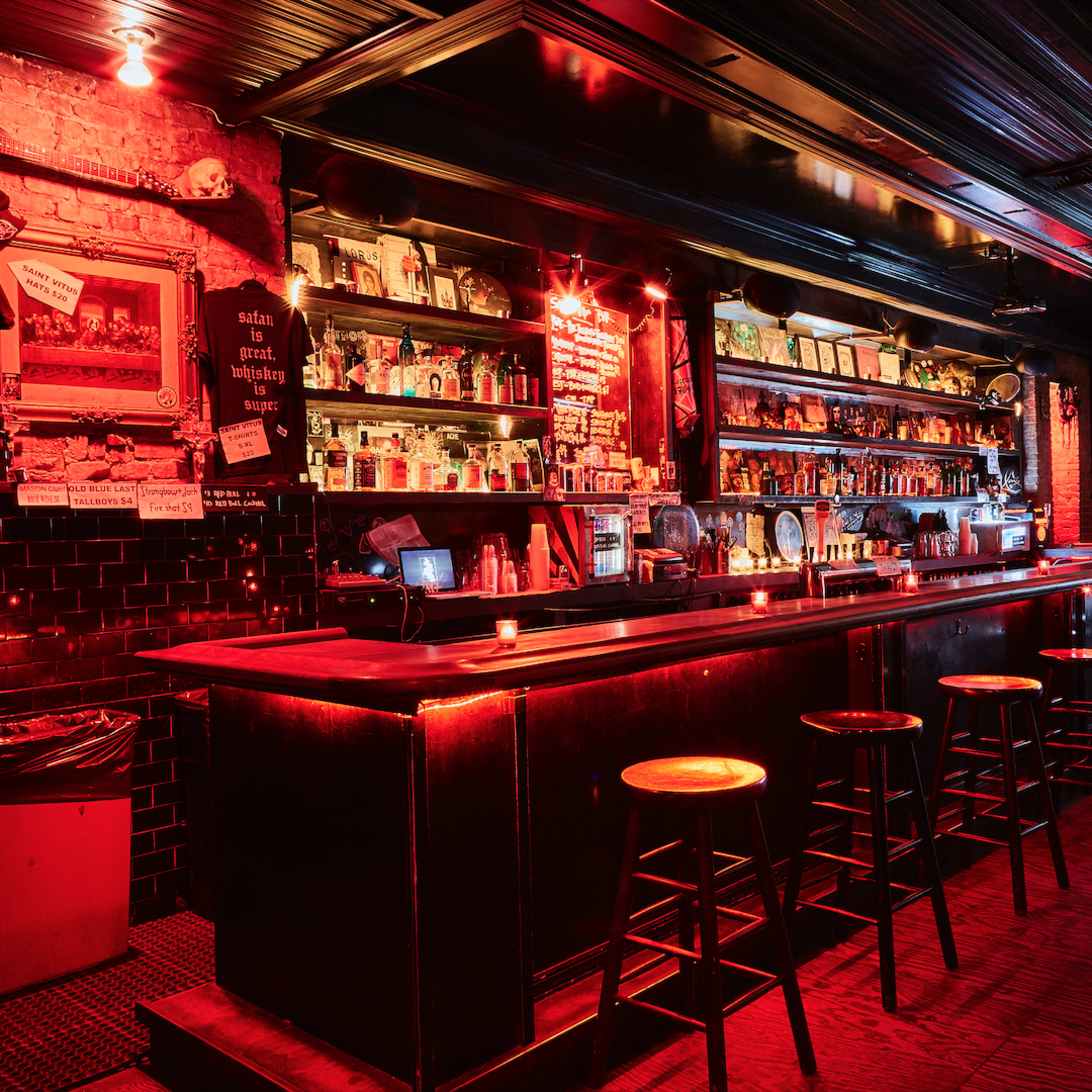 A dimly lit bar features a long dark countertop lined with stools and shelves filled with various bottles and decorations under red lighting.