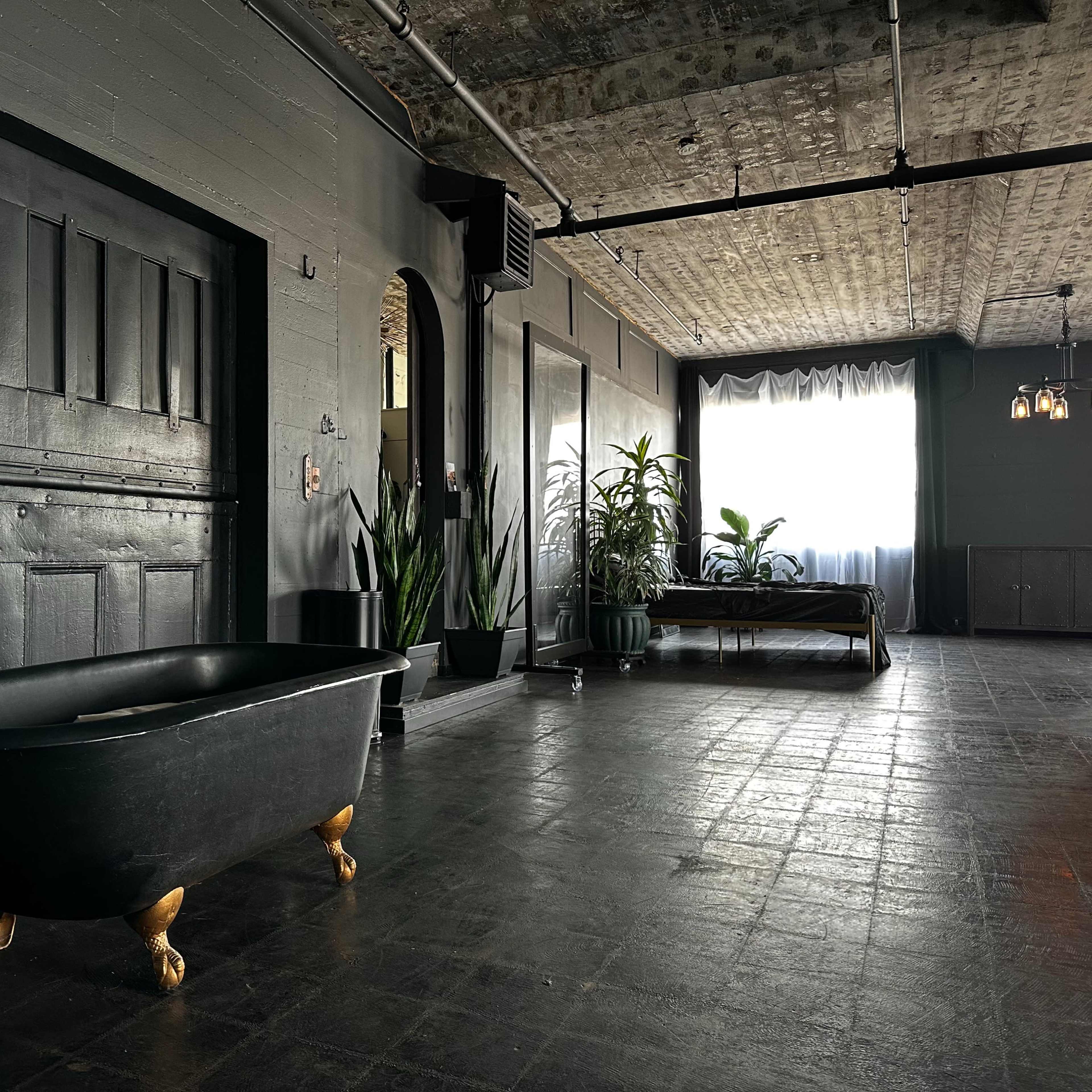 A modern interior space with a black clawfoot bathtub, large plants, and a bed positioned near a window.