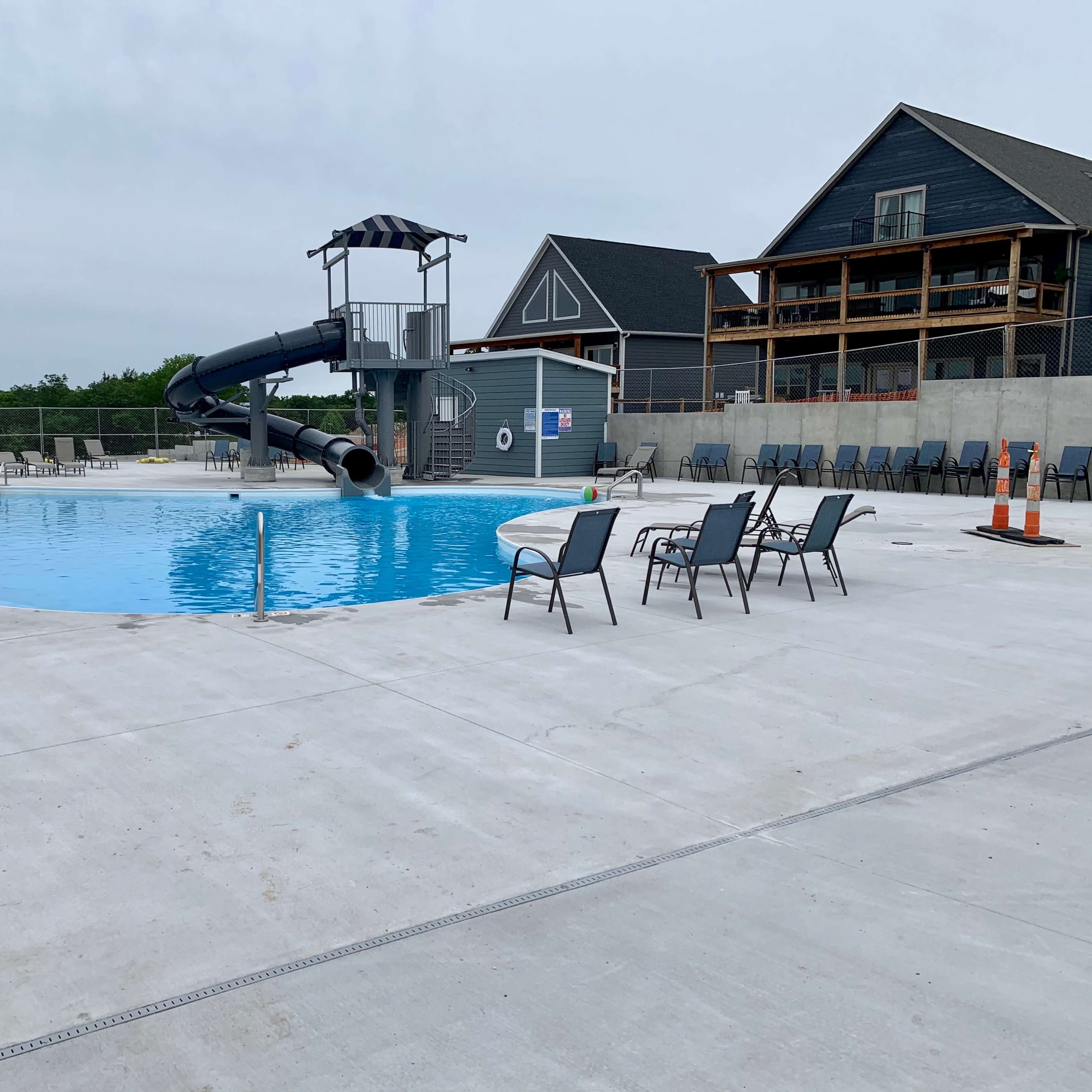 A swimming pool with a water slide, surrounded by empty lounge chairs and a building in the background.