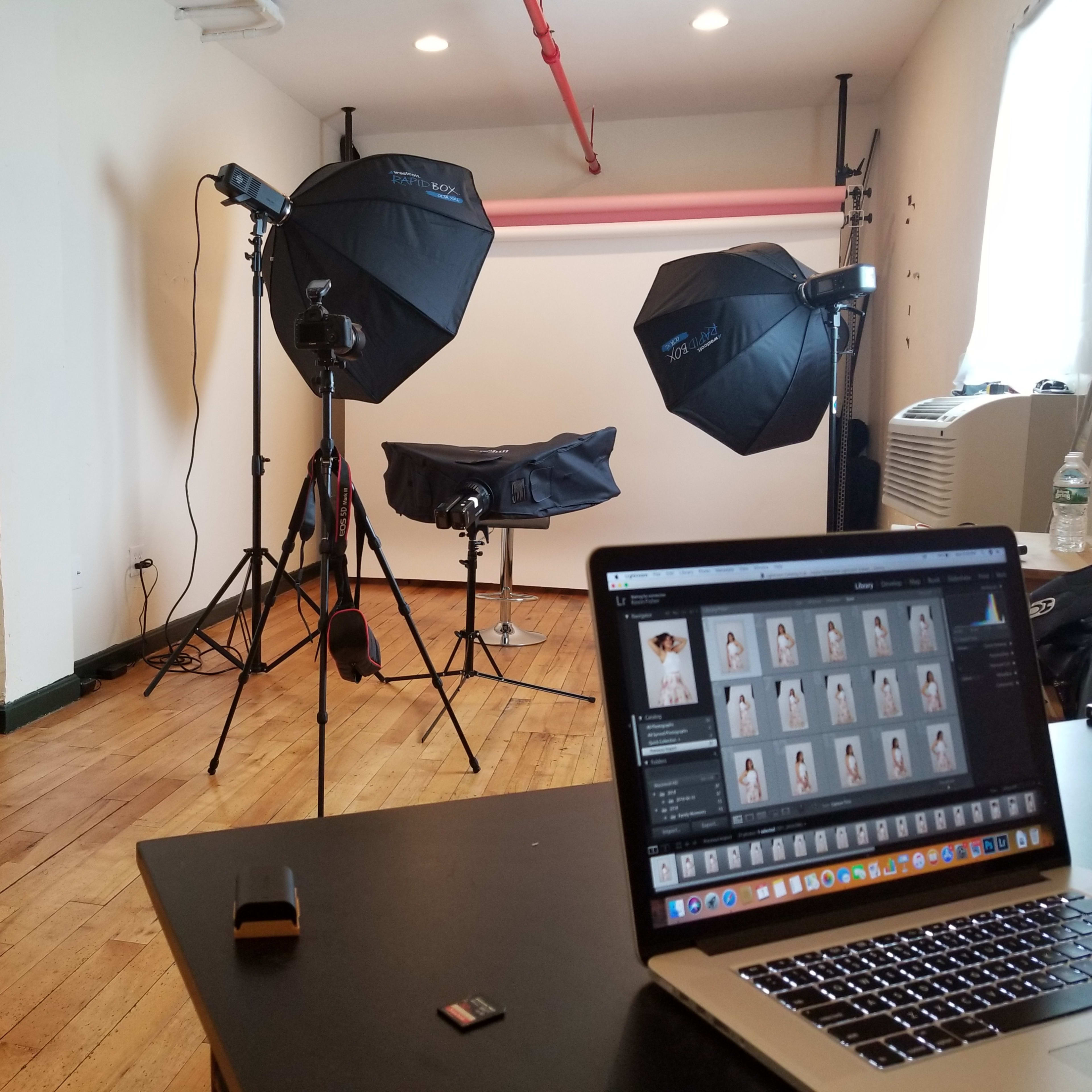 A photography studio with two softbox lights, a stool, and a computer displaying photo thumbnails on a table.
