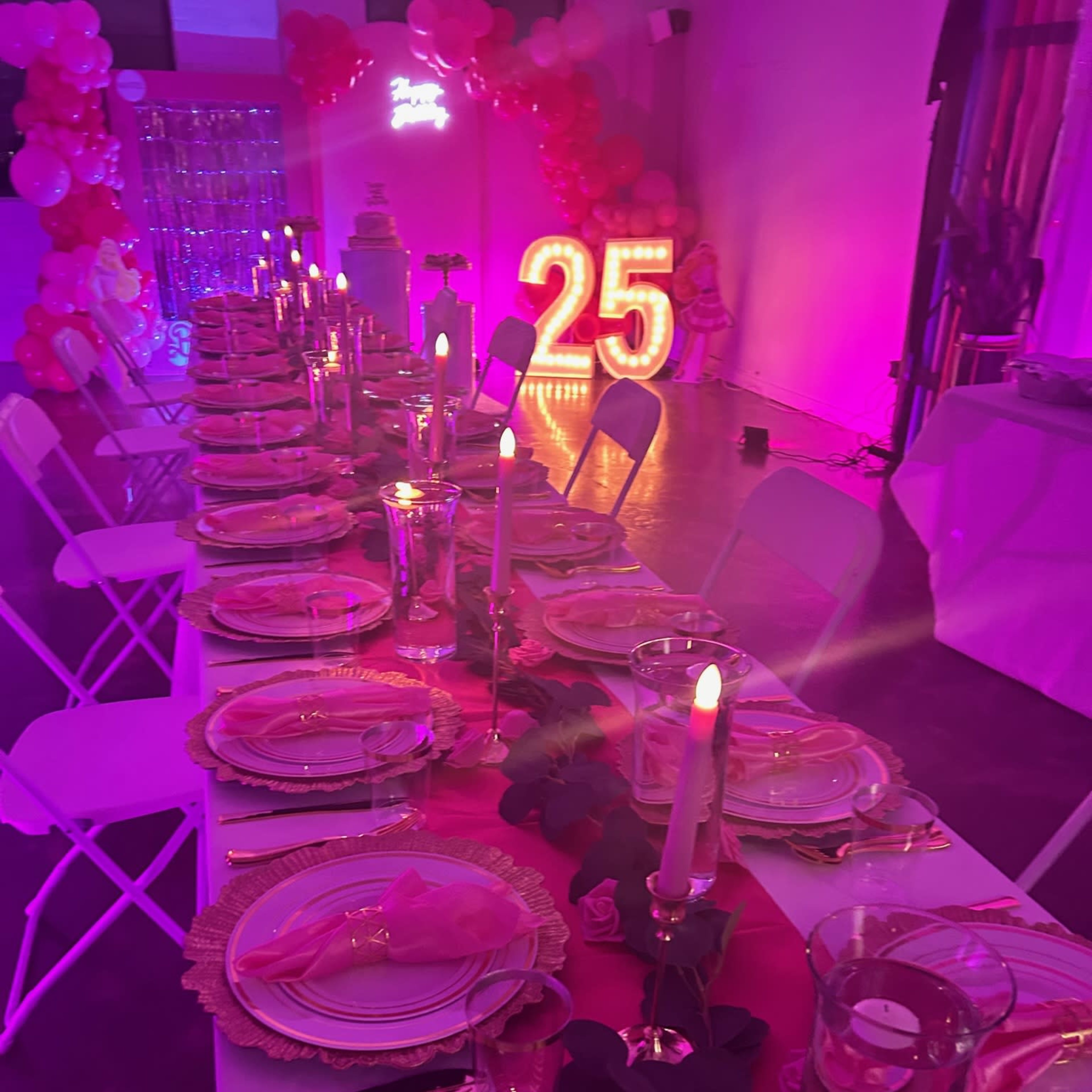 A long dining table is set with pink plates and candles, decorated with a pink runner, and illuminated by large light-up numbers "25" against a backdrop of balloons.