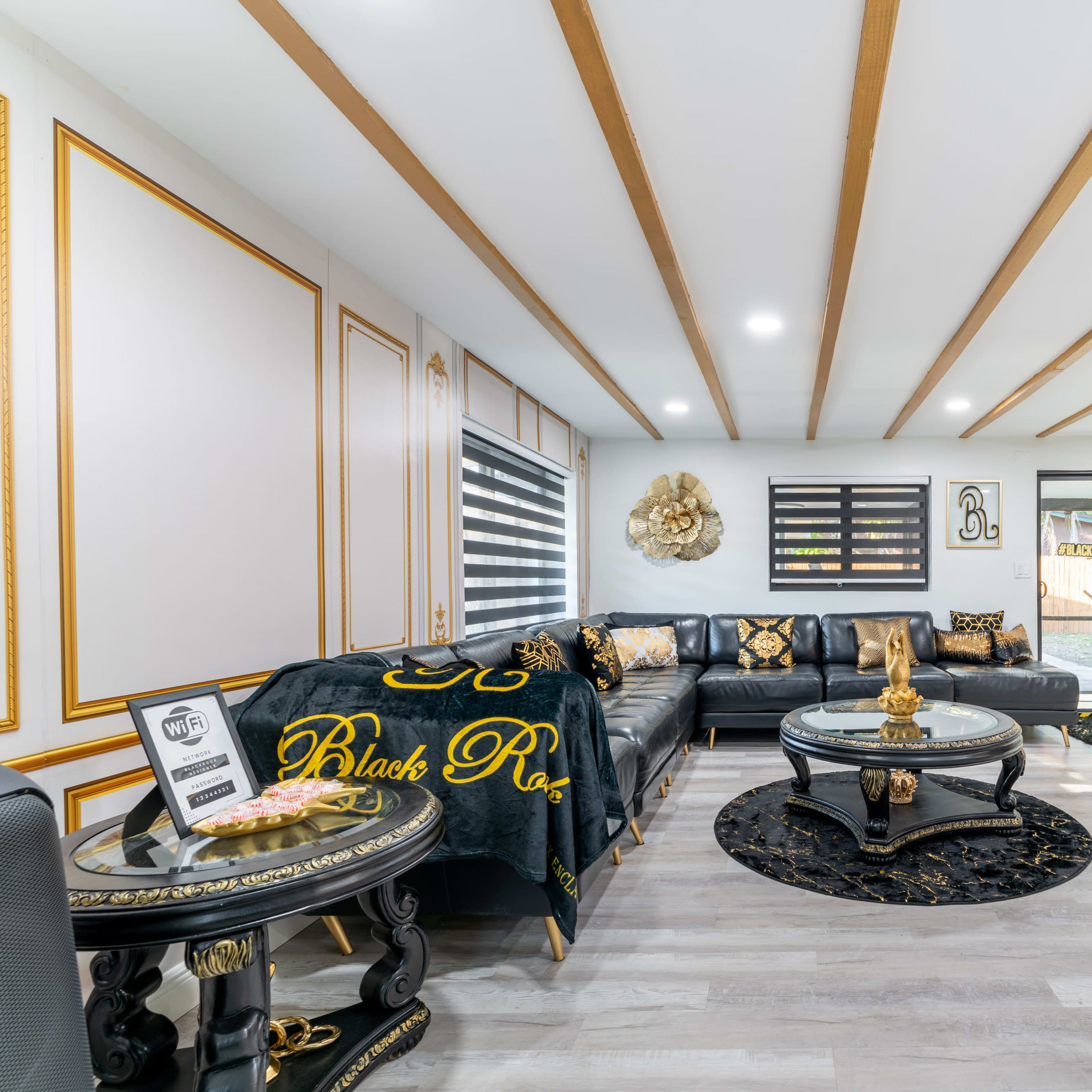 The living room features a modern design with a black leather sectional, a round glass coffee table, and decorative gold accents on the walls and furniture.