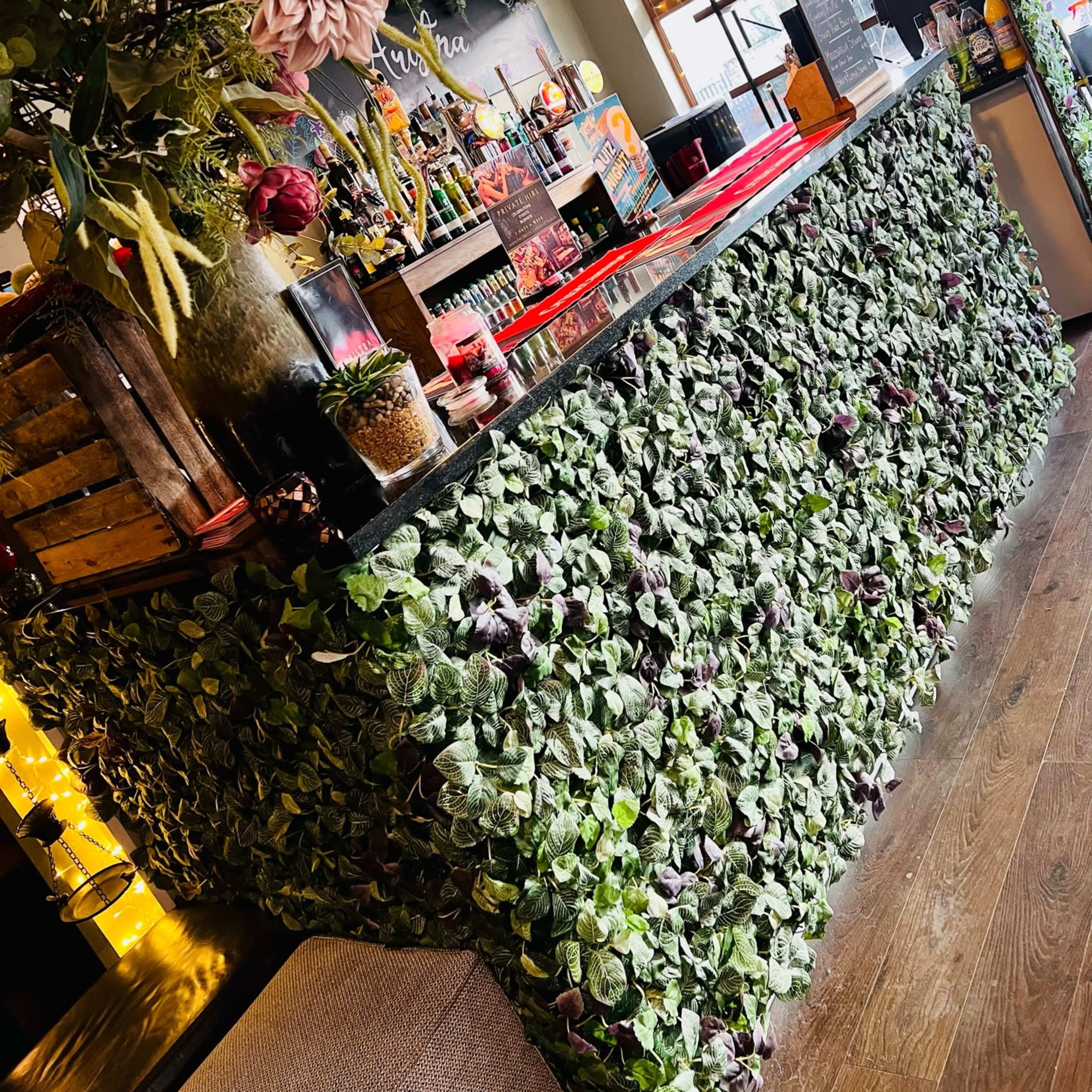 A bar counter covered in green foliage with various decorative elements and a well-stocked shelf in the background.