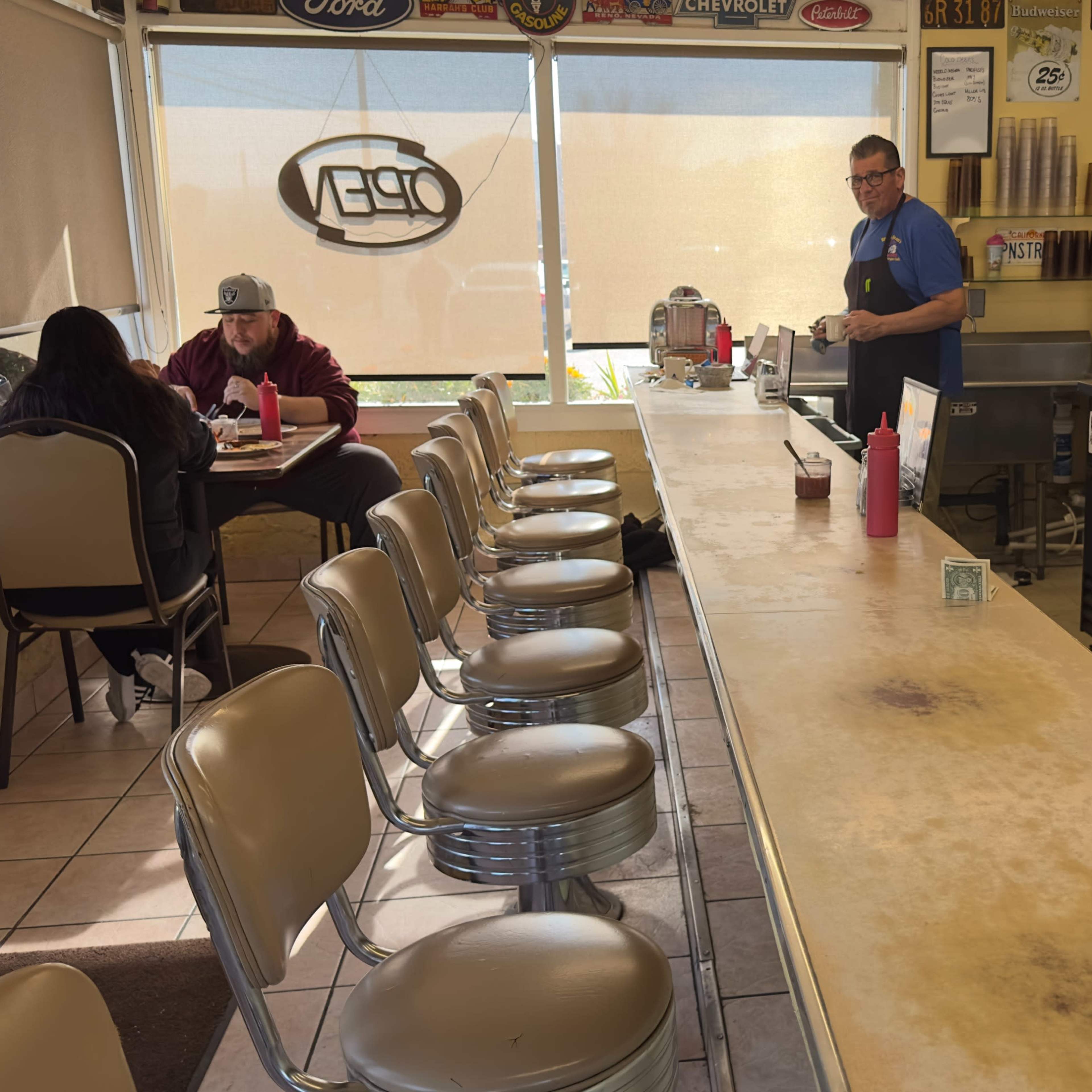 The interior of a diner with a long counter, several empty stools, and a few customers seated at tables.