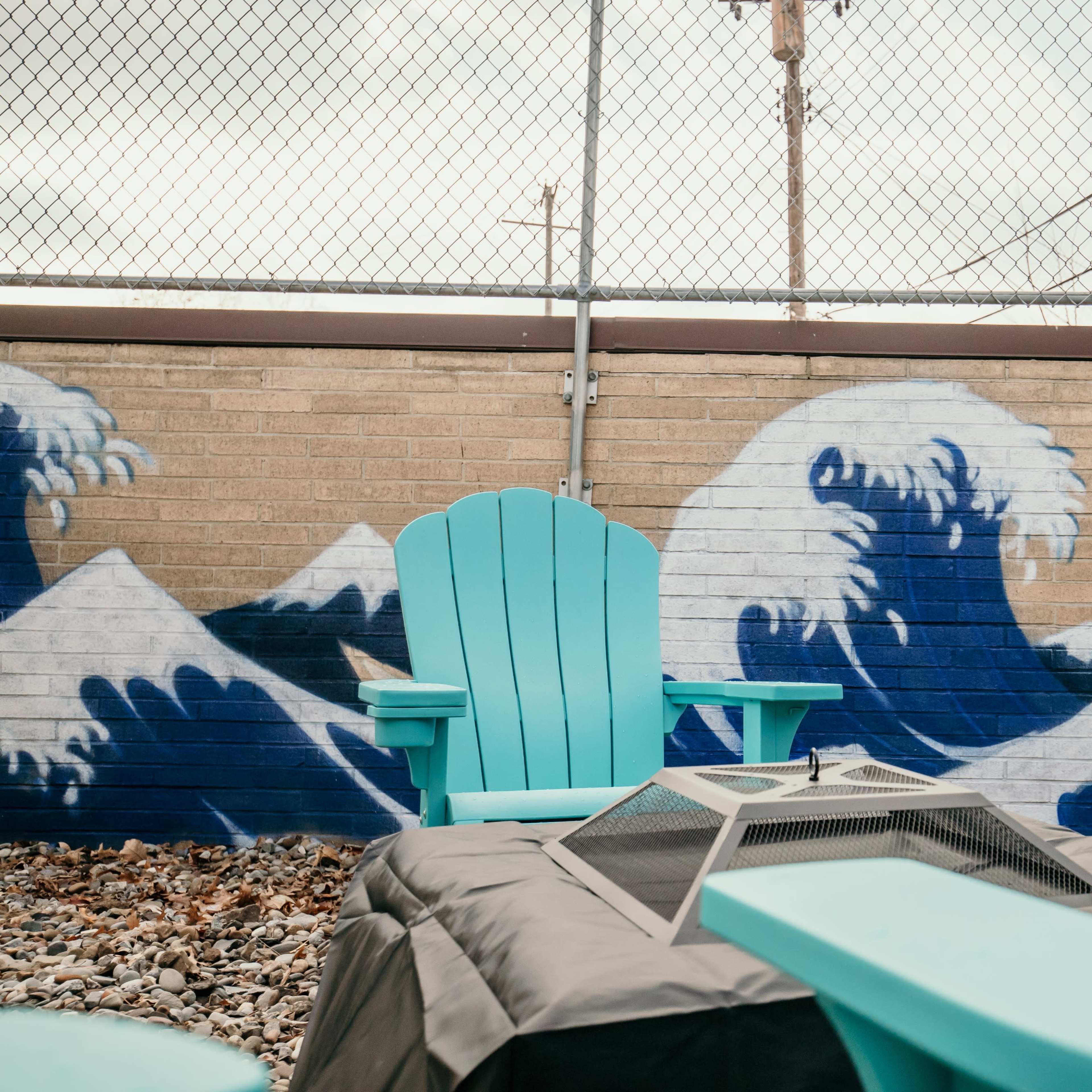 The scene features turquoise Adirondack chairs arranged around a fire pit in front of a mural depicting waves on a brick wall.