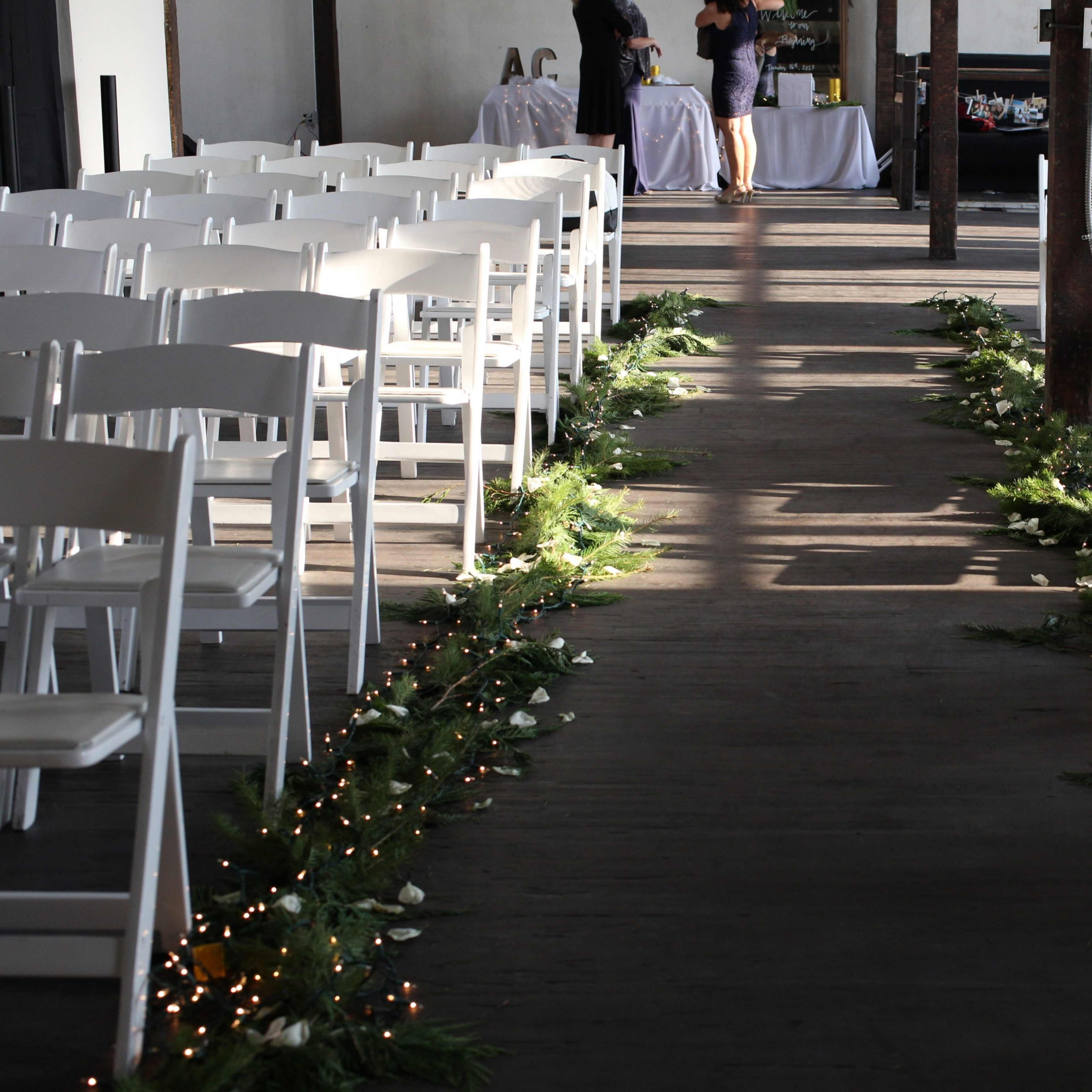A wooden aisle lined with greenery and lights, flanked by rows of white chairs set up for a ceremony.