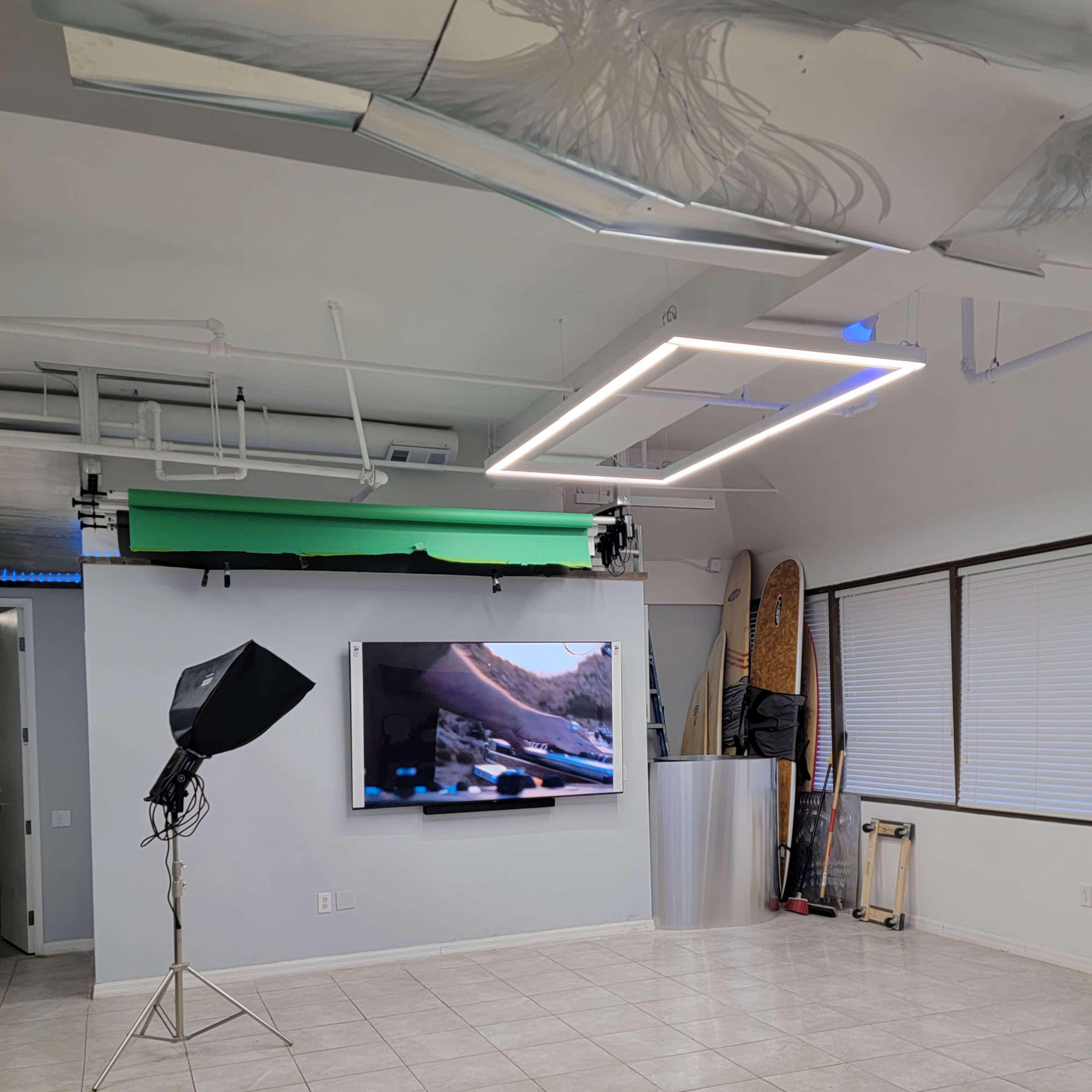 A bright room equipped with a green screen setup, a large television on the wall, and surfboards leaning against the side.