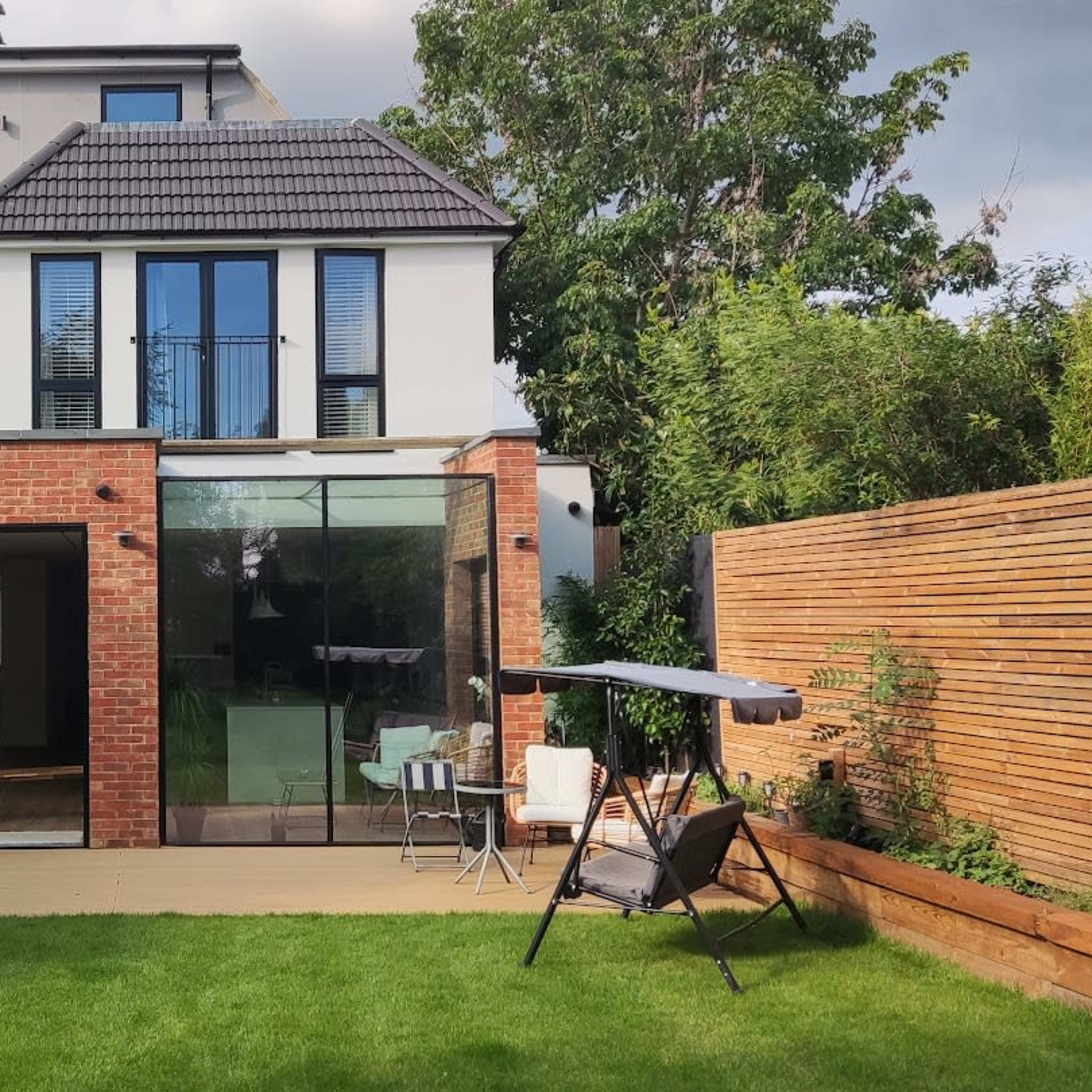 A modern house with a brick and glass extension, surrounded by a well-maintained garden featuring a wooden deck and outdoor seating.