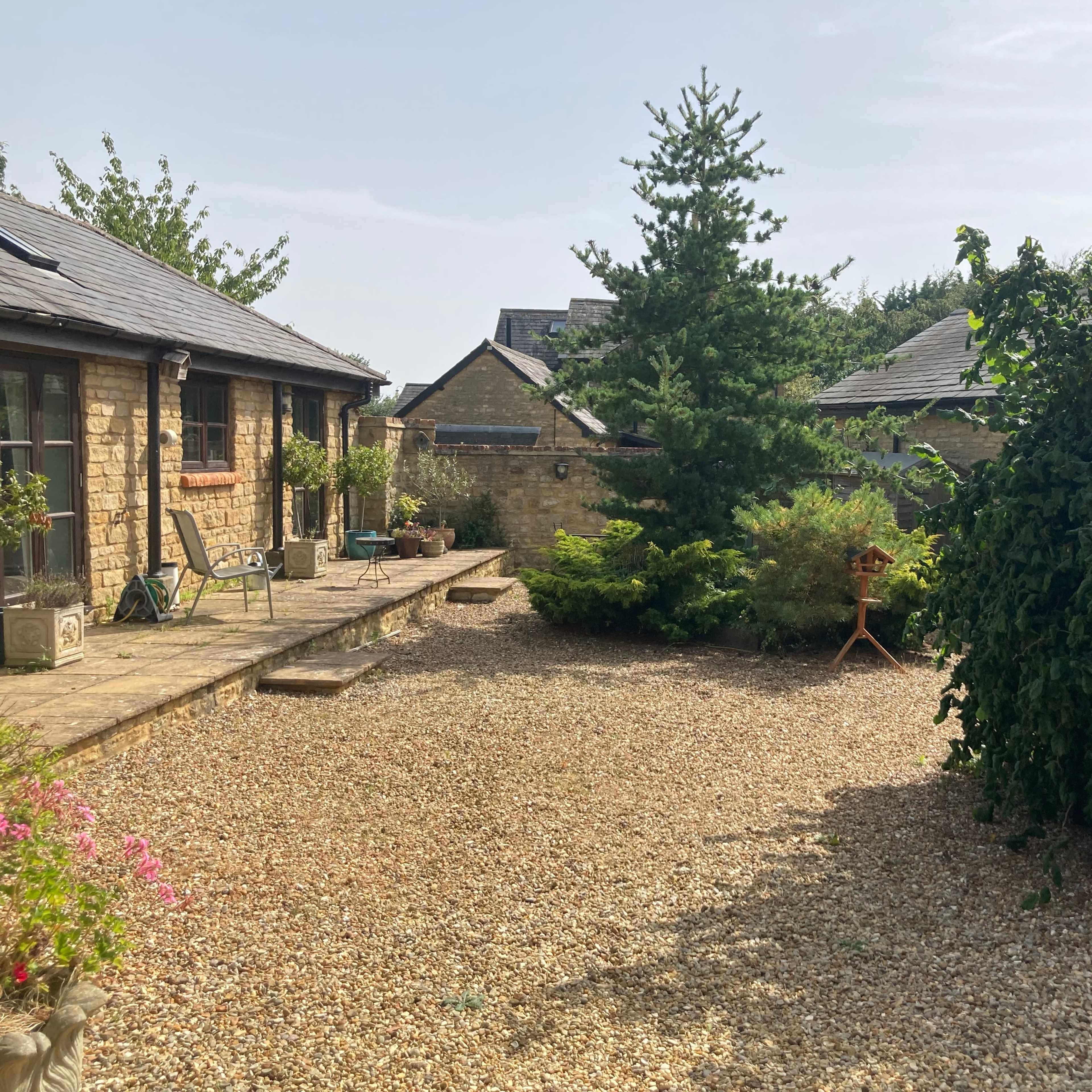 A gravel area surrounded by stone buildings and greenery, including a large bush and a pine tree.