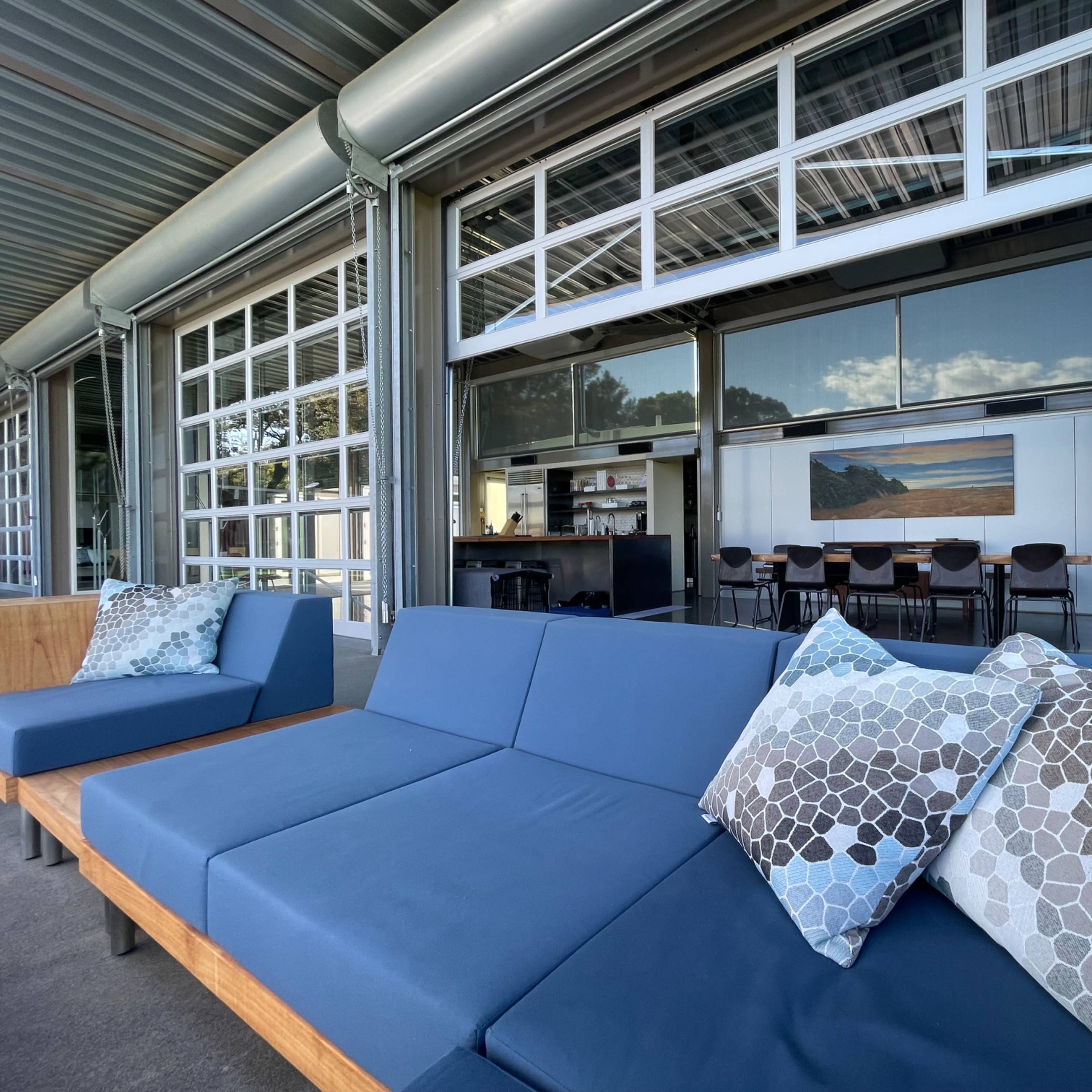 A modern outdoor seating area with a blue sofa adorned with patterned pillows, overlooking an open space that includes a kitchen and dining area behind large glass doors.