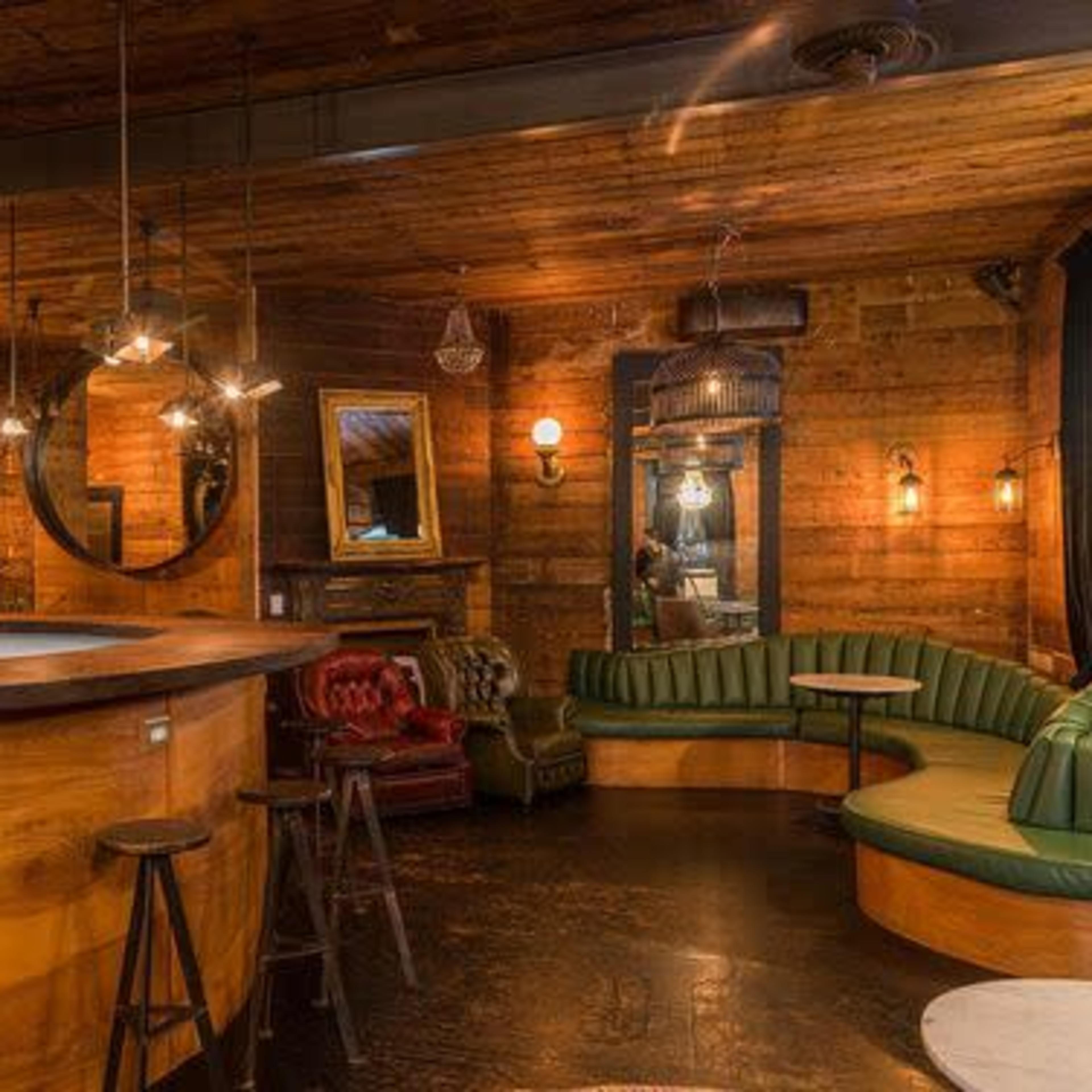 A dimly lit bar interior featuring wooden walls, circular mirrors, and a mix of seating arrangements including a curved green leather bench and red armchairs.