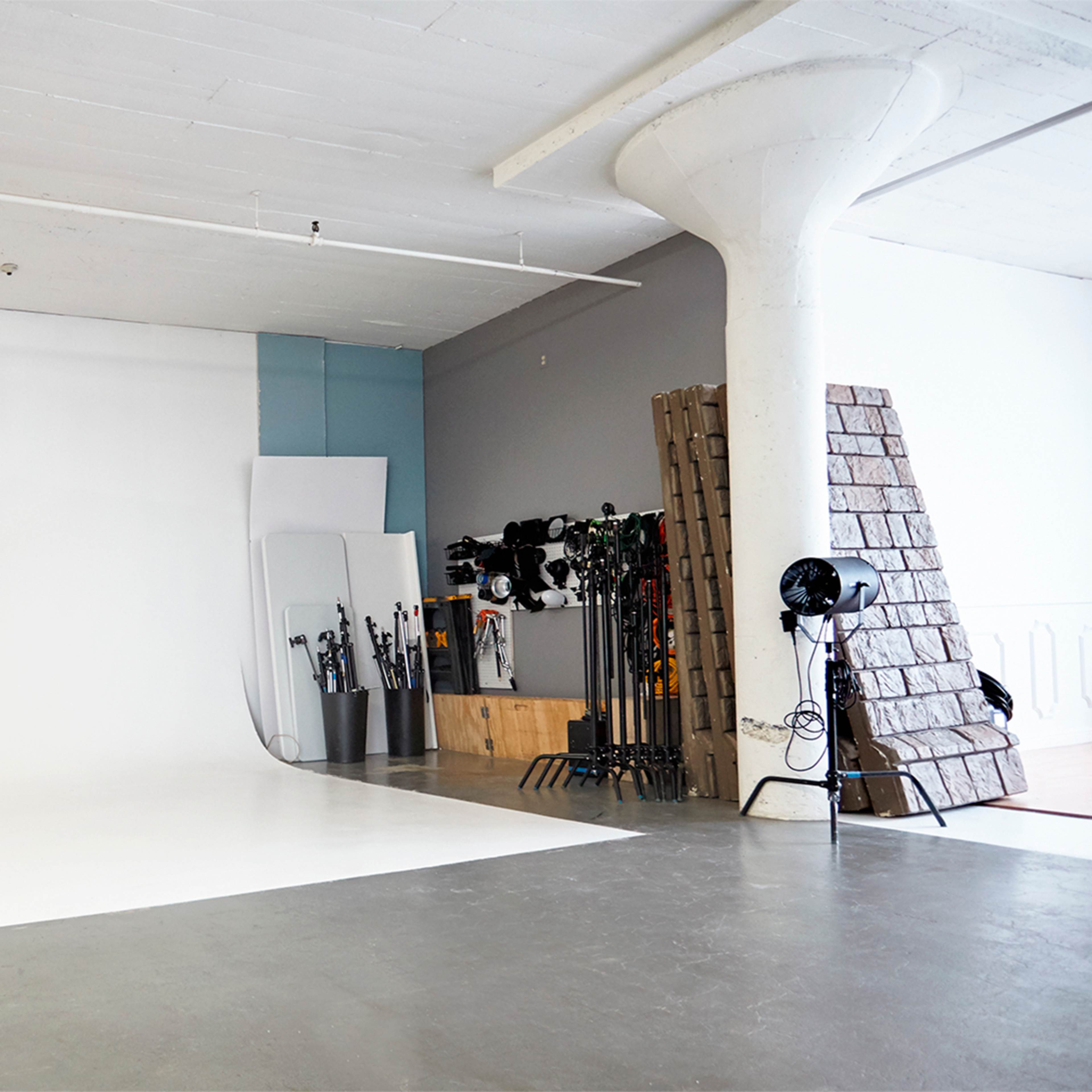 A spacious photography studio with a white backdrop, a brick wall, and various lighting equipment arranged against the walls.