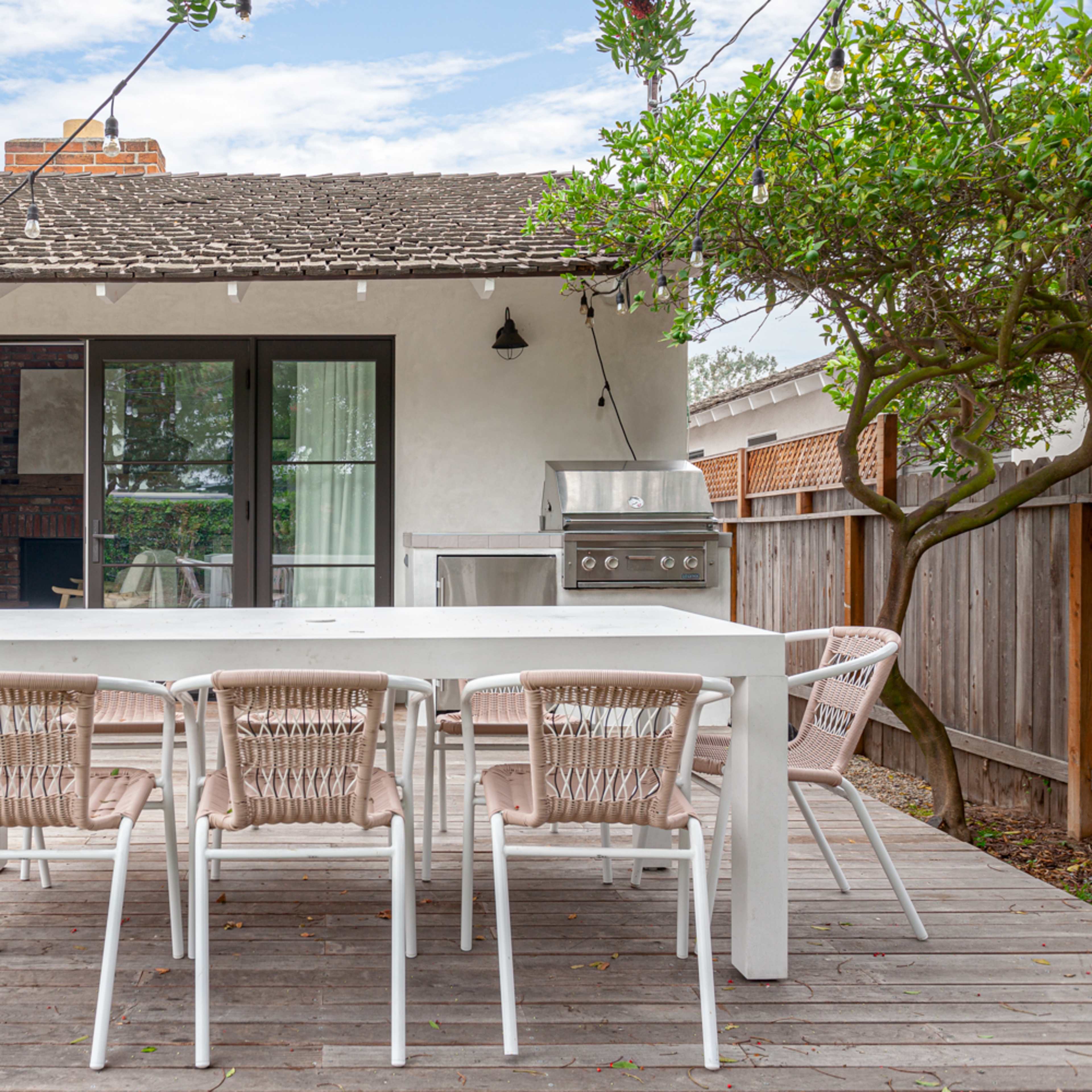 A modern outdoor dining area with a white table and matching chairs, situated on a wooden deck surrounded by greenery and a barbecue grill.