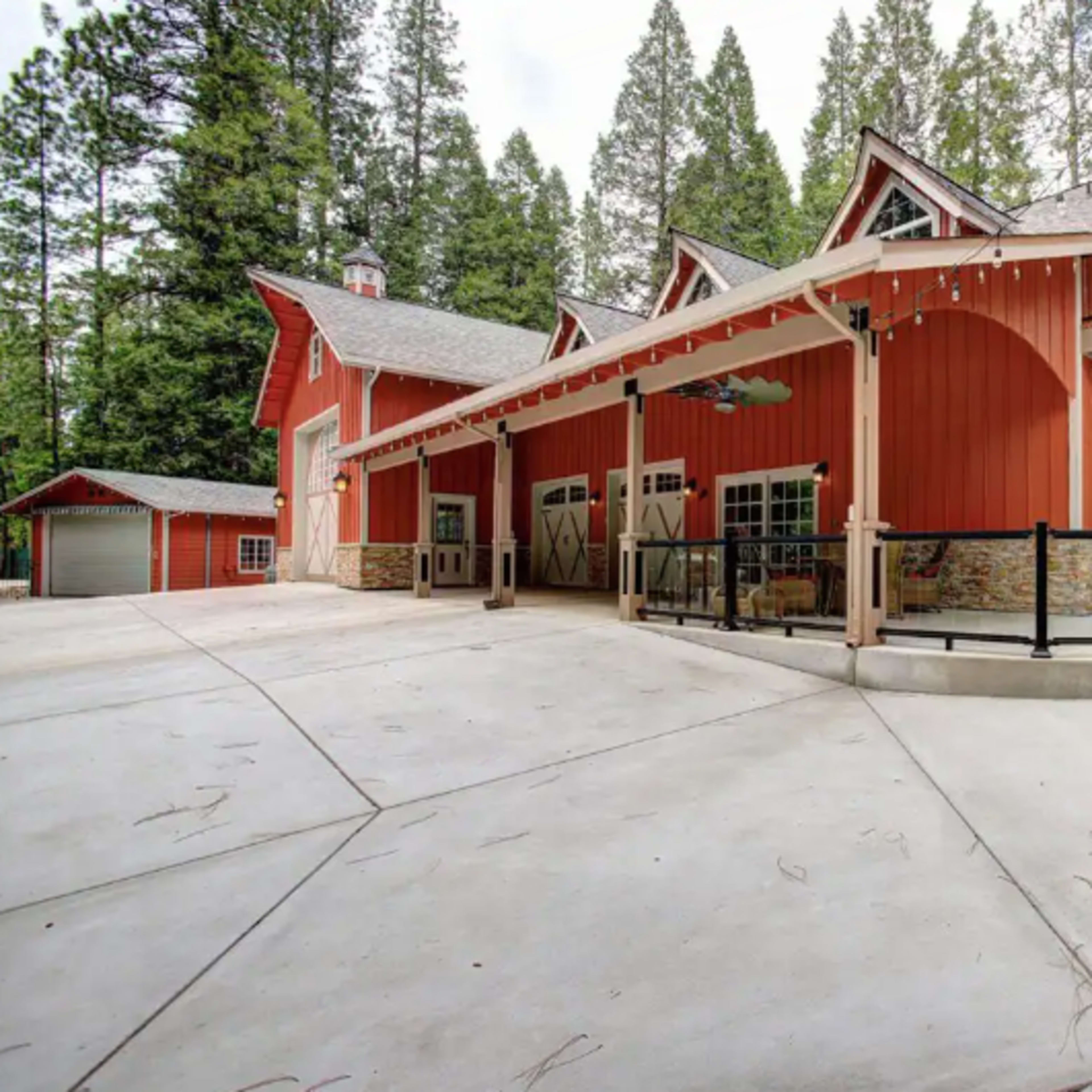 A red and gray multi-level house with a wraparound porch, surrounded by tall green trees.