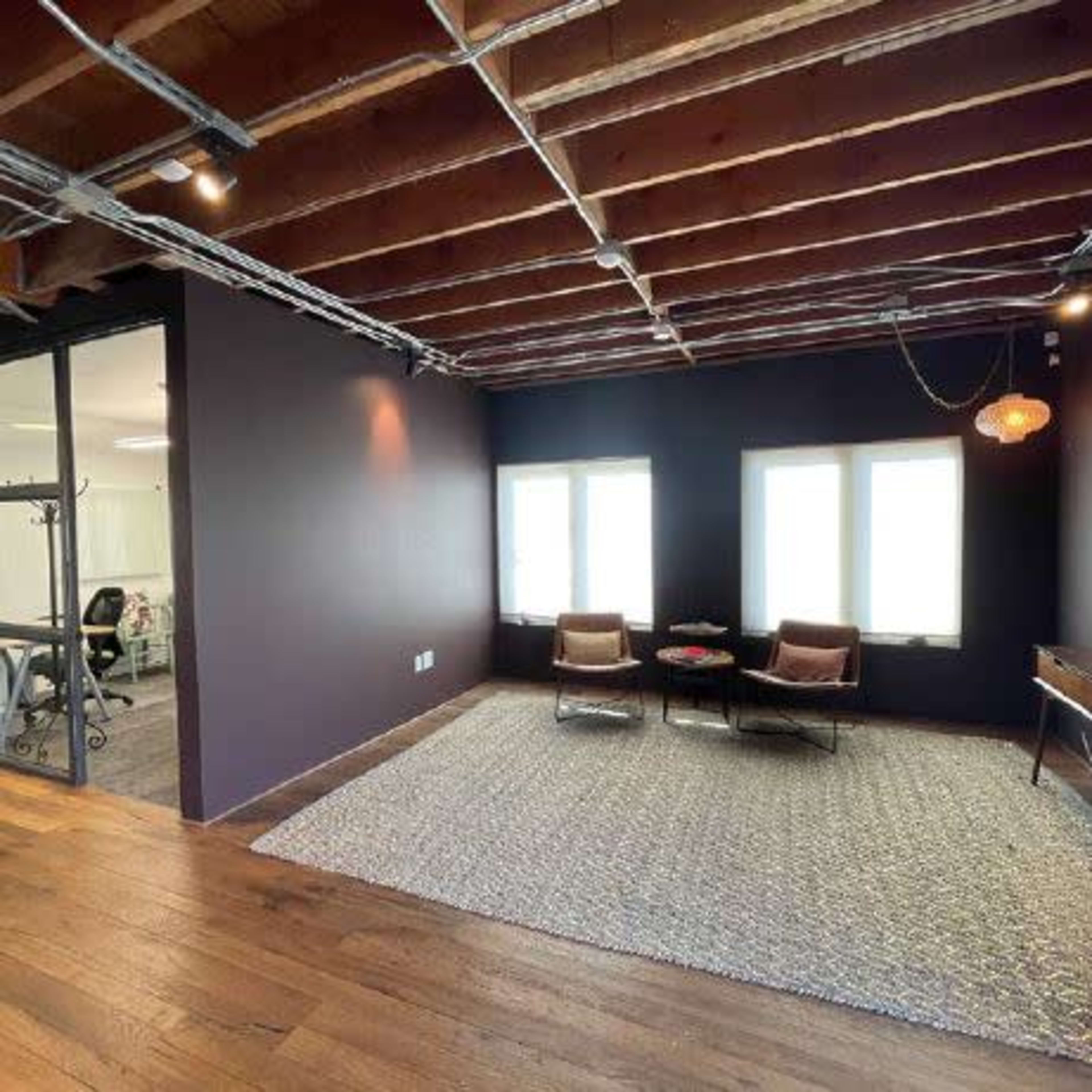 A modern office space with dark walls, wooden beams, a rug, and a seating area featuring two chairs and a coffee table near large windows.