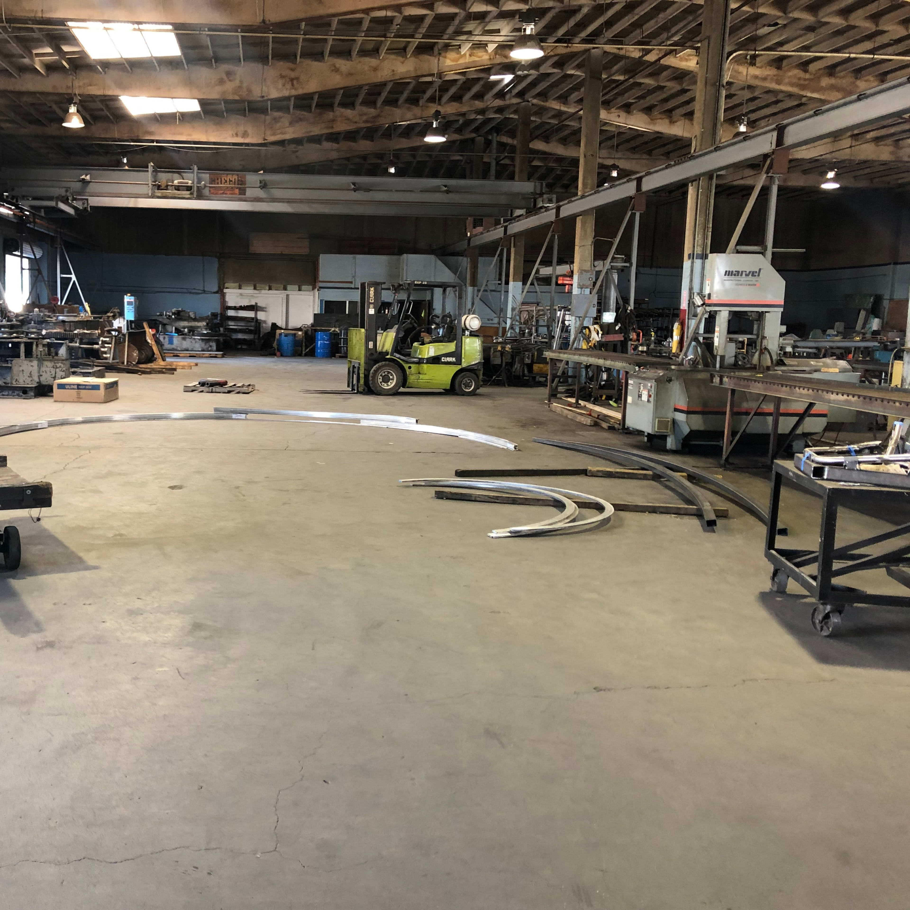 A spacious industrial workshop with machinery, a forklift, and metal materials scattered across the concrete floor.