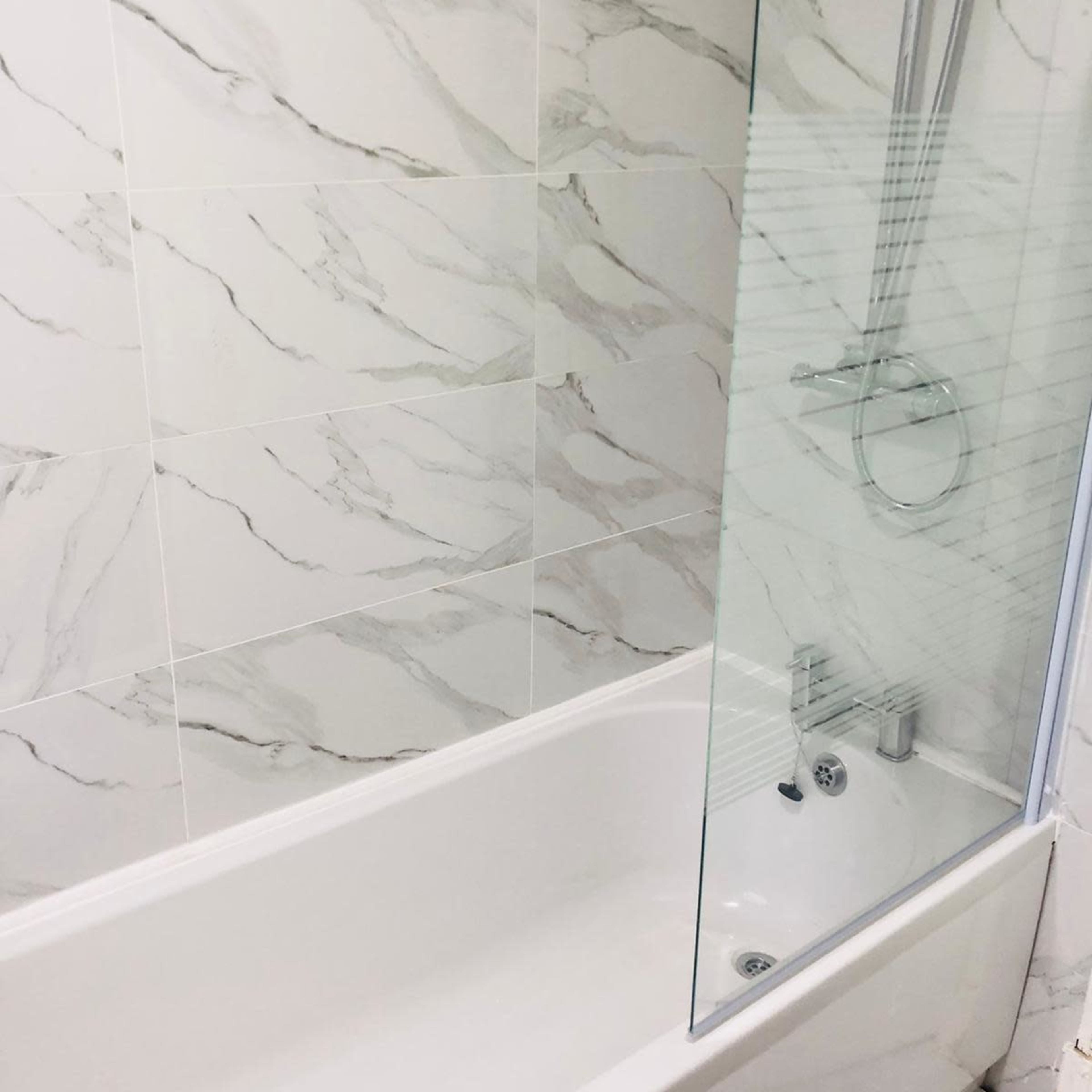 A modern bathroom featuring a white tub with a glass shower screen and marble-patterned tiled walls.