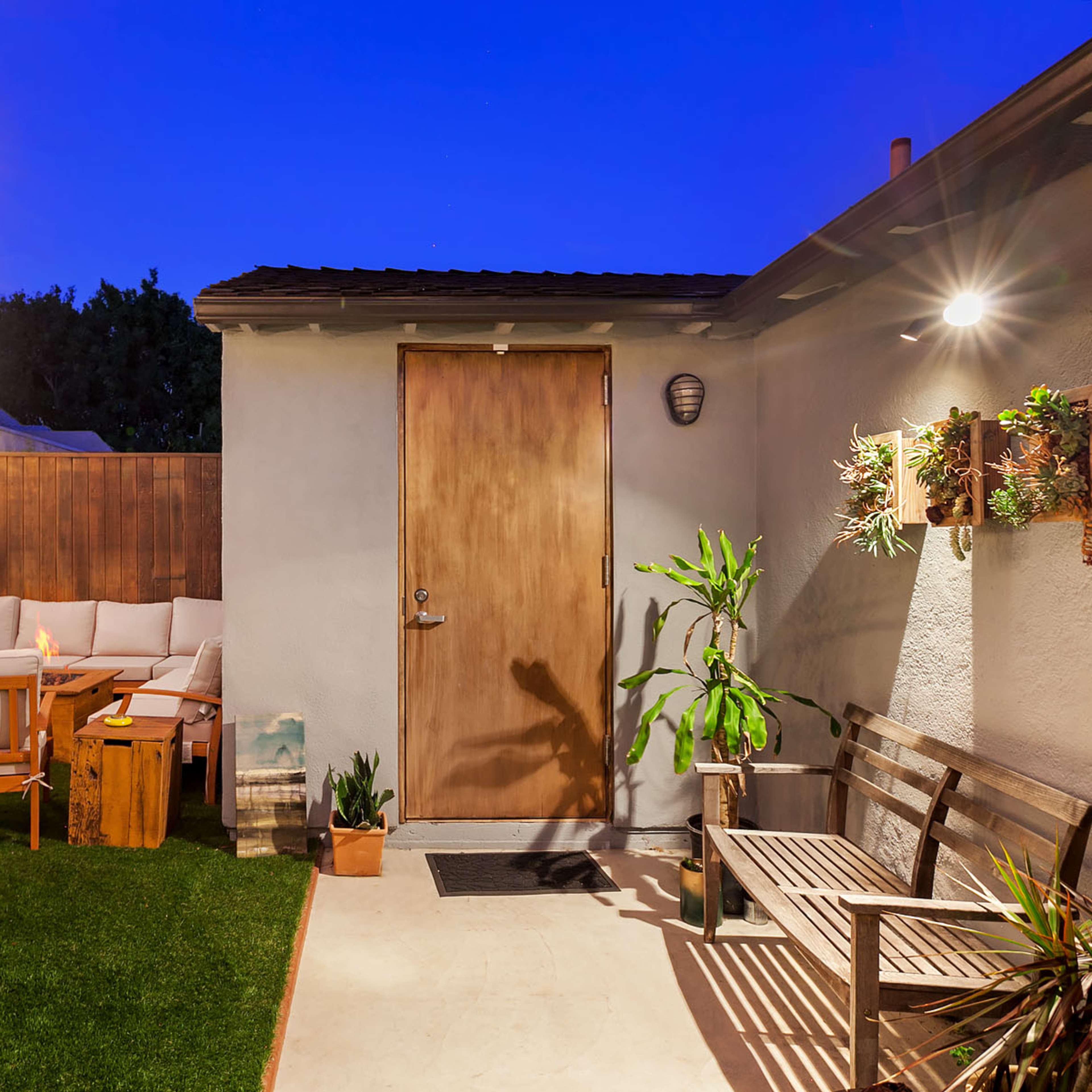 A backyard area featuring a wooden door, outdoor seating arrangements, decorative plants, and string lights against a twilight sky.