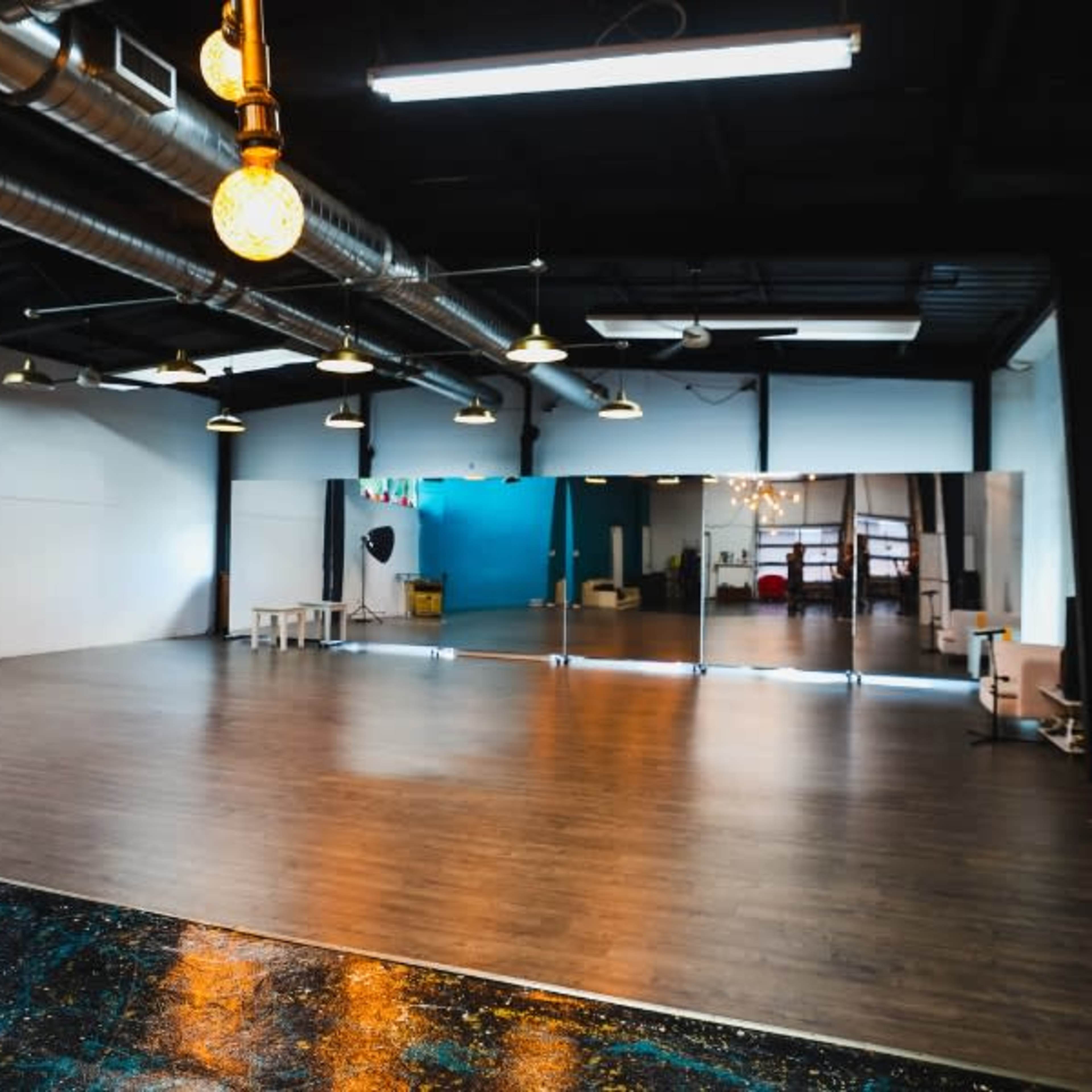An empty studio space with wooden flooring, large mirrors on one wall, and industrial lighting fixtures.
