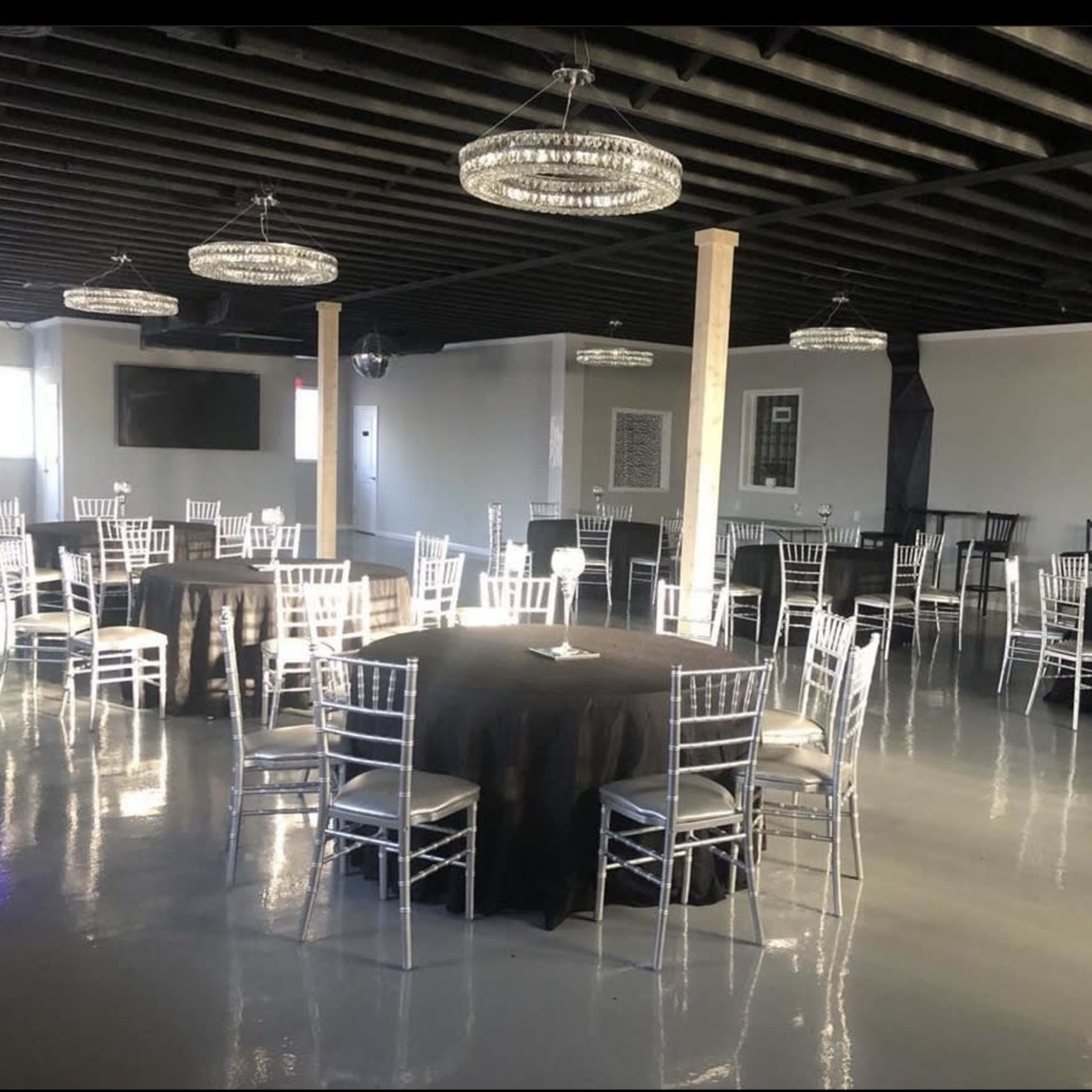 A spacious event venue with black round tables surrounded by silver chiavari chairs and modern chandelier lighting.