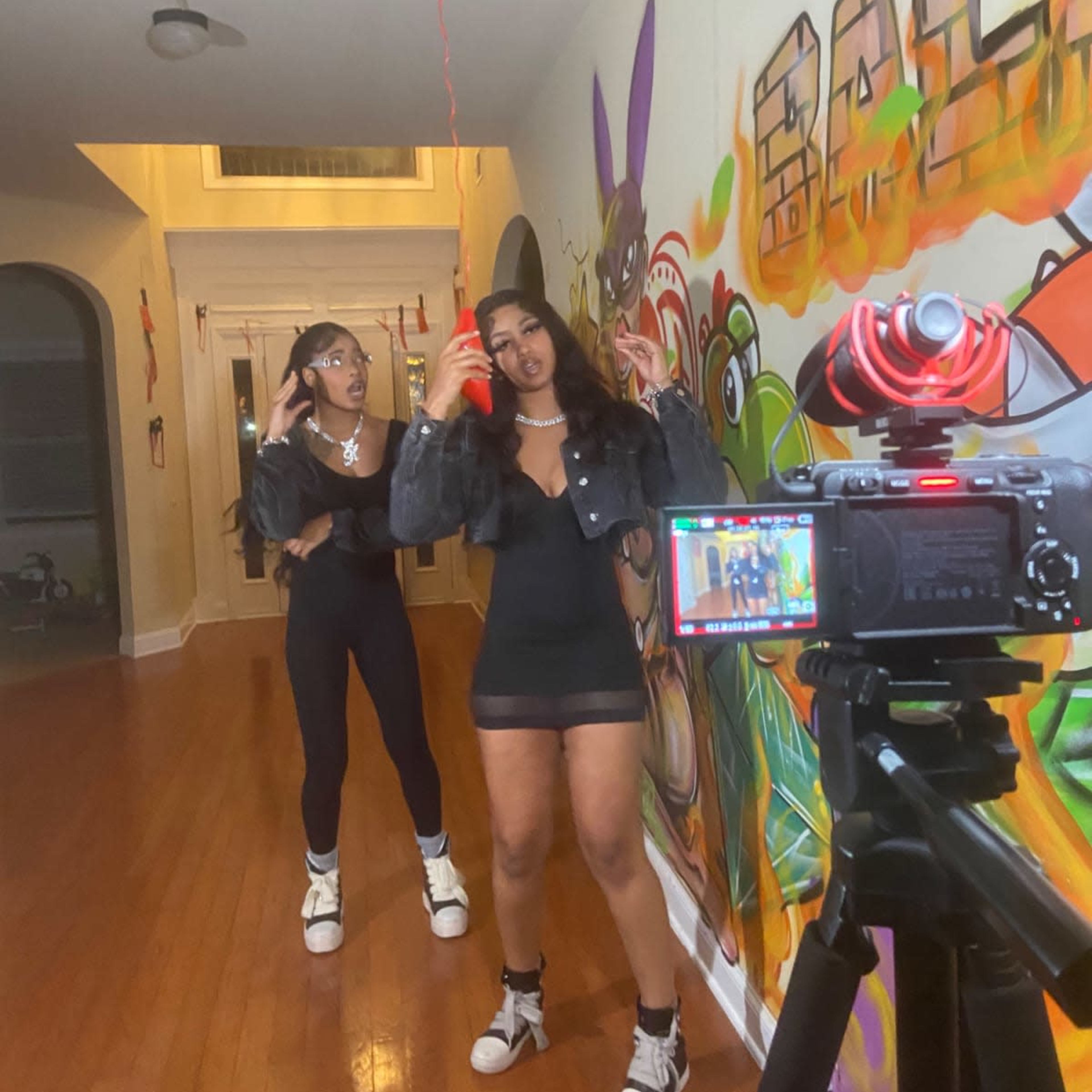 Two young women pose for a photo in a hallway adorned with colorful graffiti, while a camera on a tripod captures the scene.